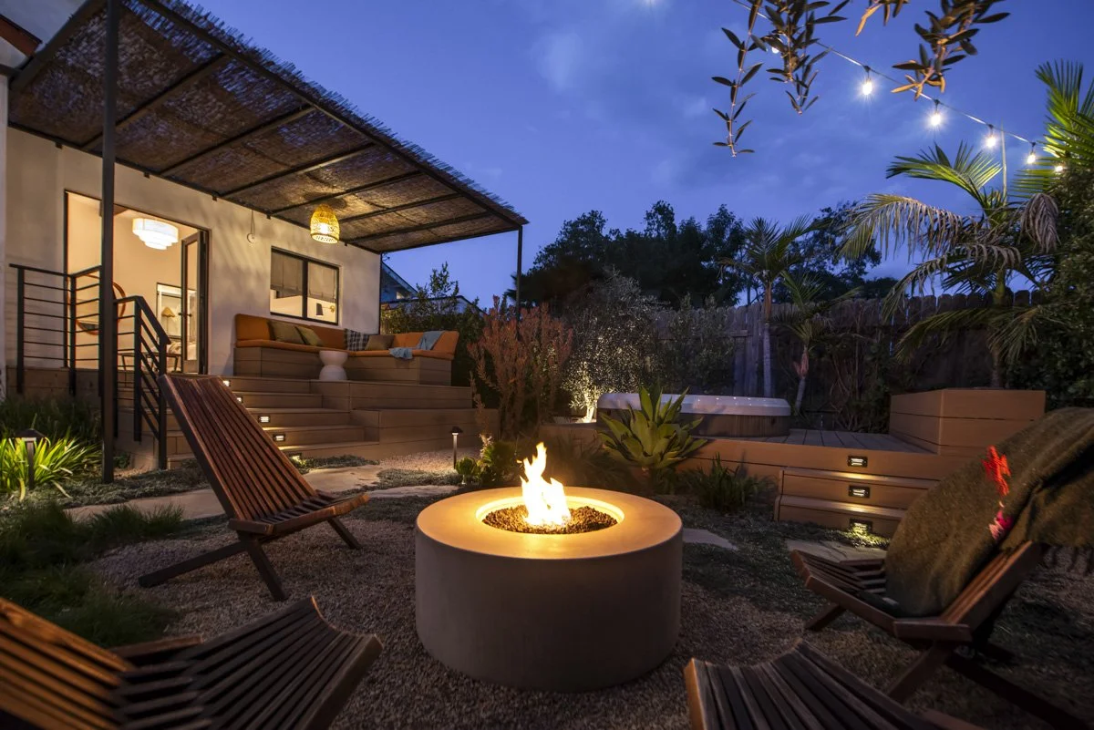 A cozy backyard patio at dusk with a fire pit, wooden lounge chairs, a hot tub, and string lights overhead, surrounded by lush plants and trees.