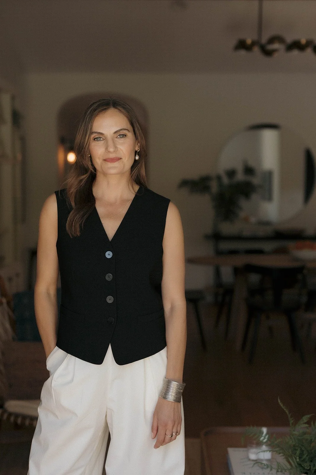 Woman wearing a black sleeveless vest and white pants standing in a room with a dining area in the background.