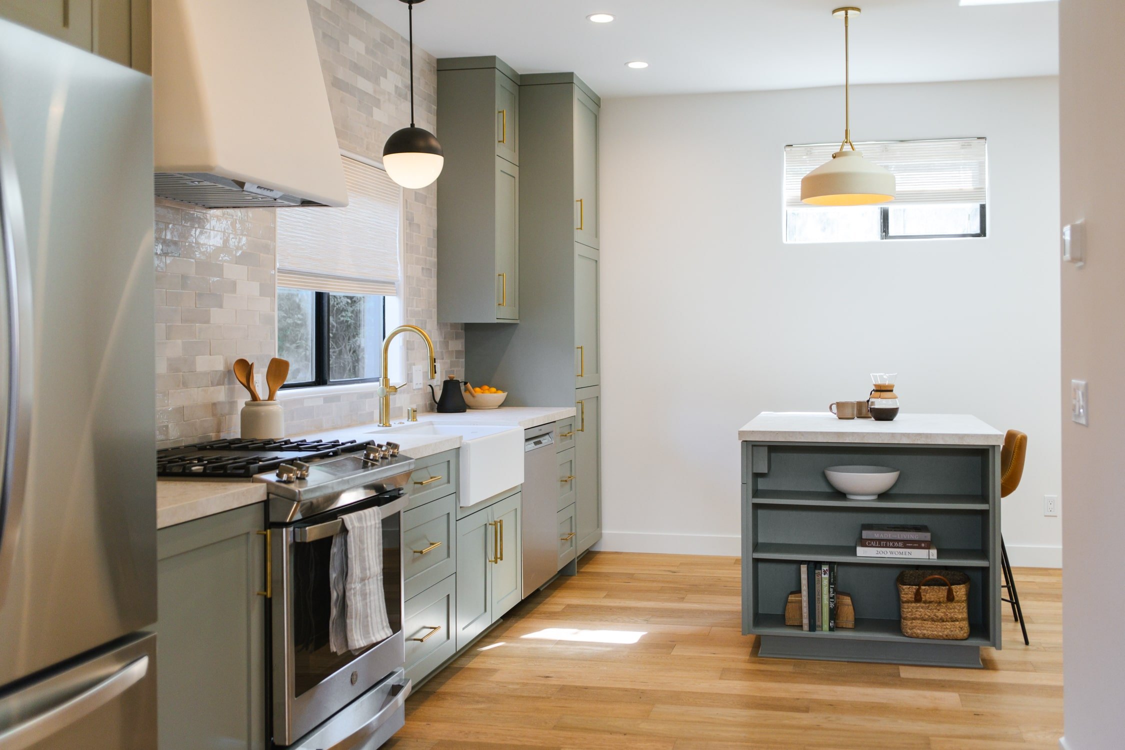 A modern kitchen with gray cabinets, a white farmhouse sink, a stainless steel stove, hanging pendant lights, a small island with open shelving, a window with blinds, and hardwood floors.