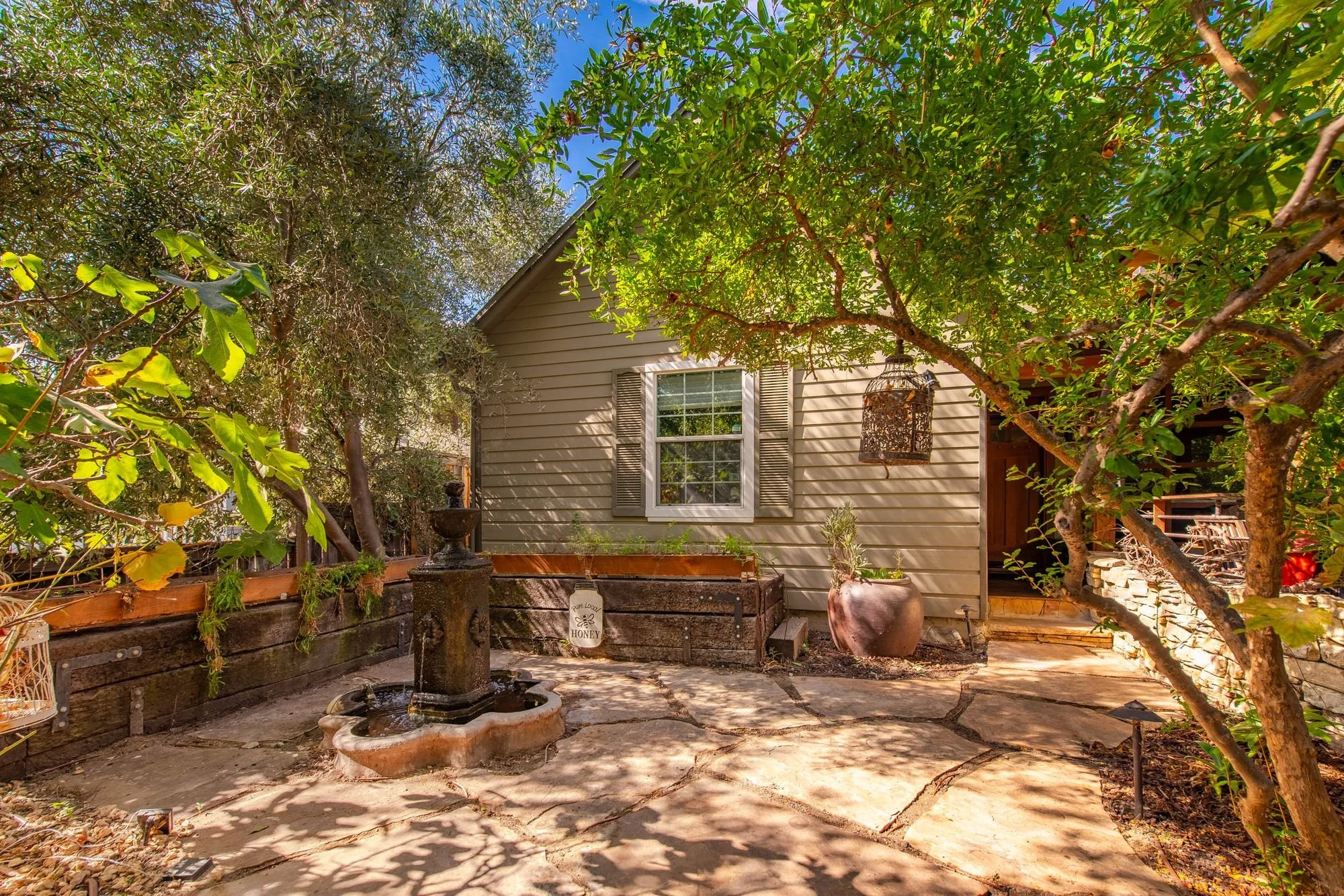 A backyard garden with a small fountain, several potted plants, trees, and a house with a window, surrounded by a wooden fence and lush green foliage.