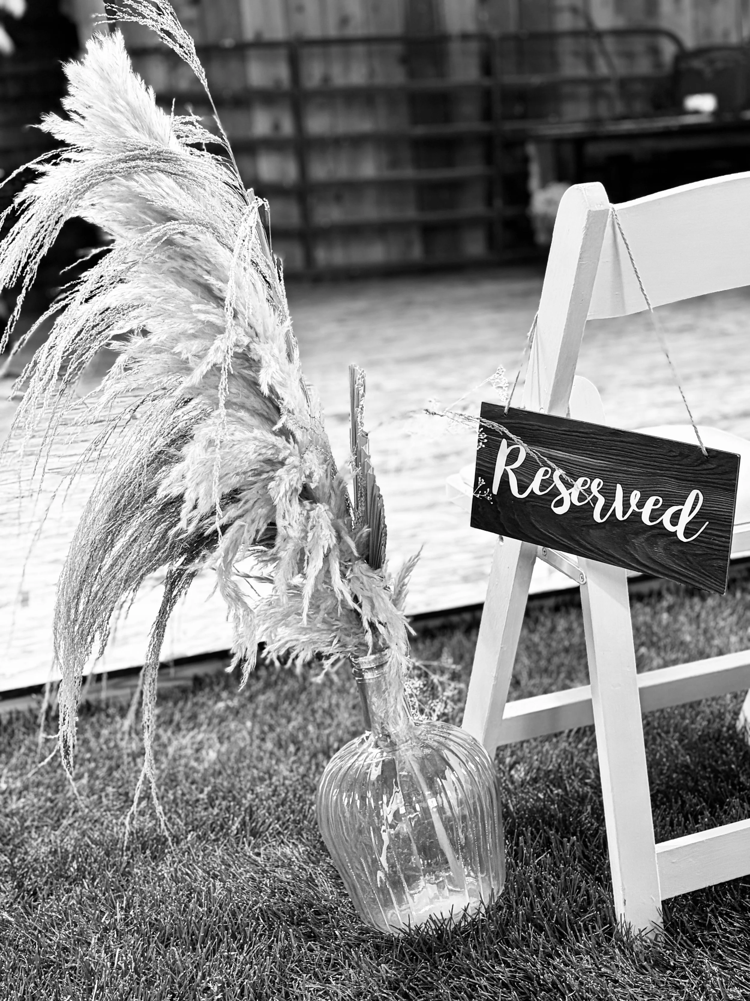 A glass vase with pampas grass and dried ornamental grass, placed next to a small white wooden chair with a reserved sign hanging on it, on a grassy surface.