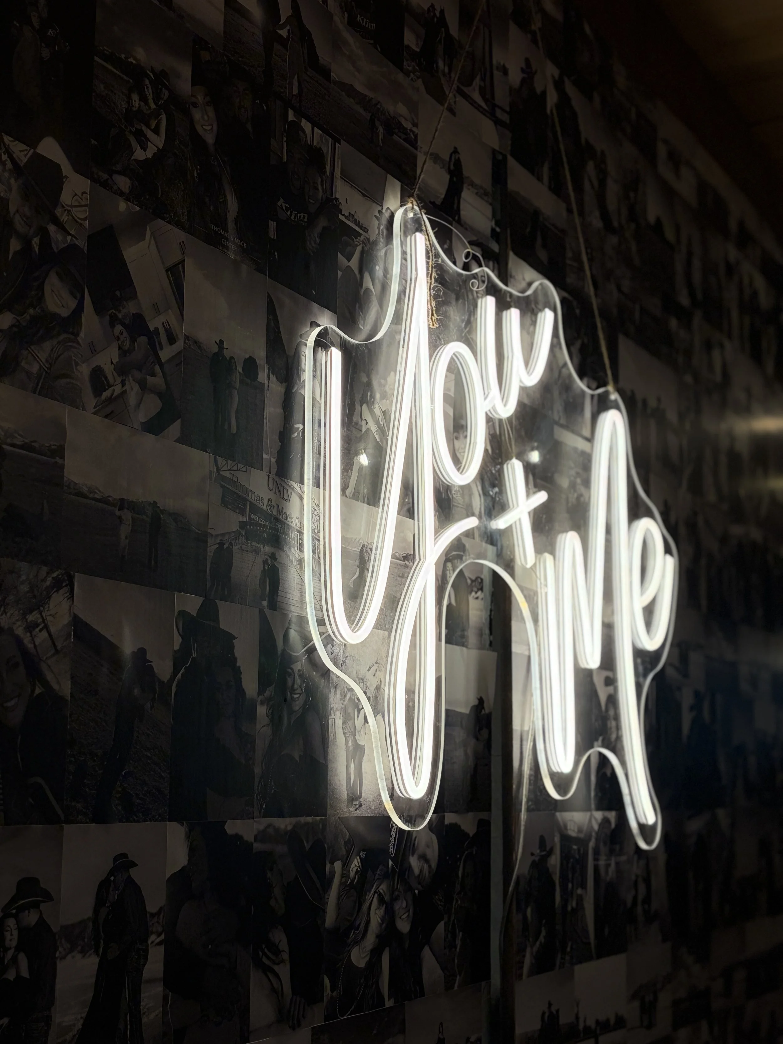 Close-up of a neon sign with a cursive word, hanging in front of a collage of black-and-white photographs of people, including some in cowboy hats and outdoor scenes, on a dark wall.