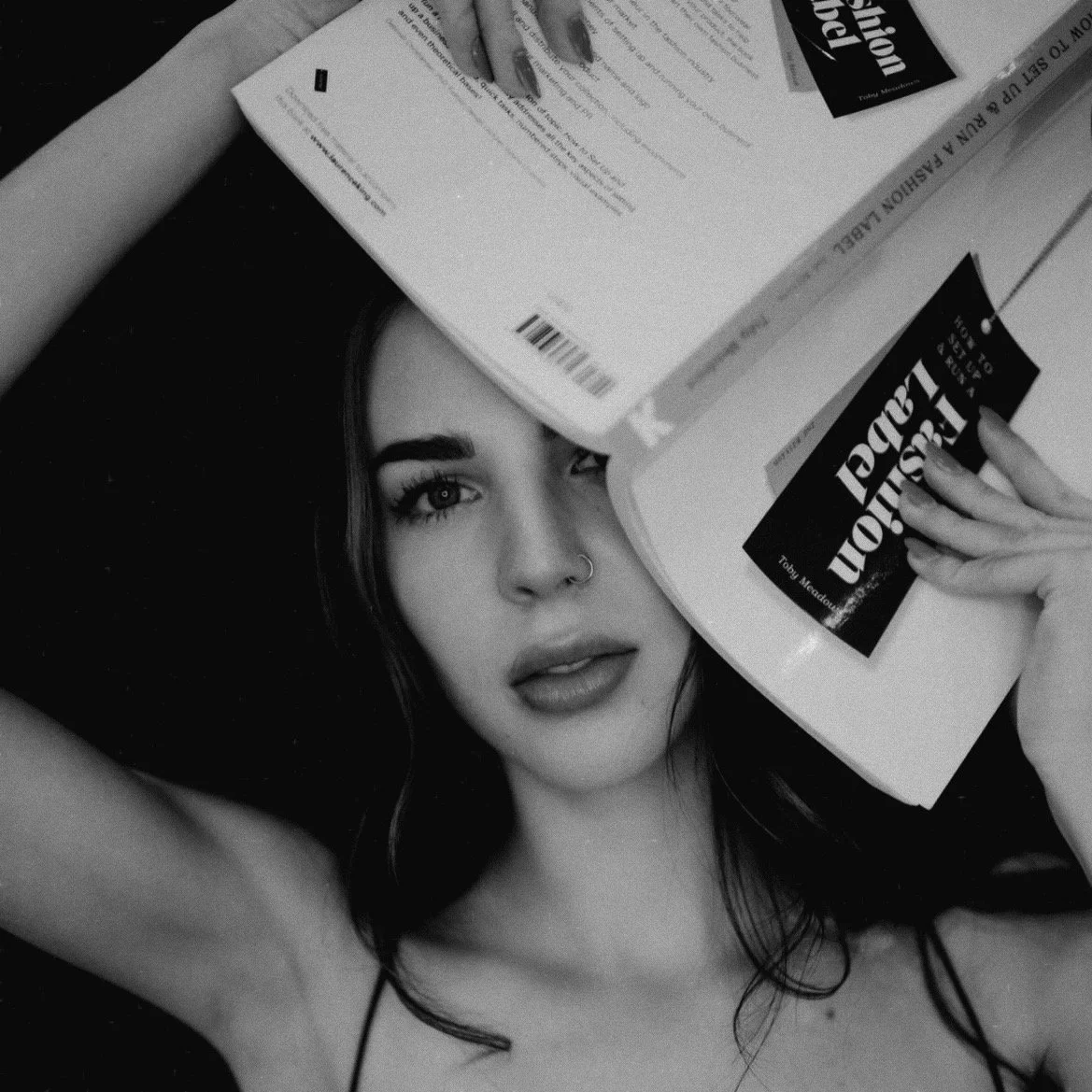 Black and white photo of a woman with dark hair and makeup holding an open book with the title "FASHION LOGIC" covering part of her face.