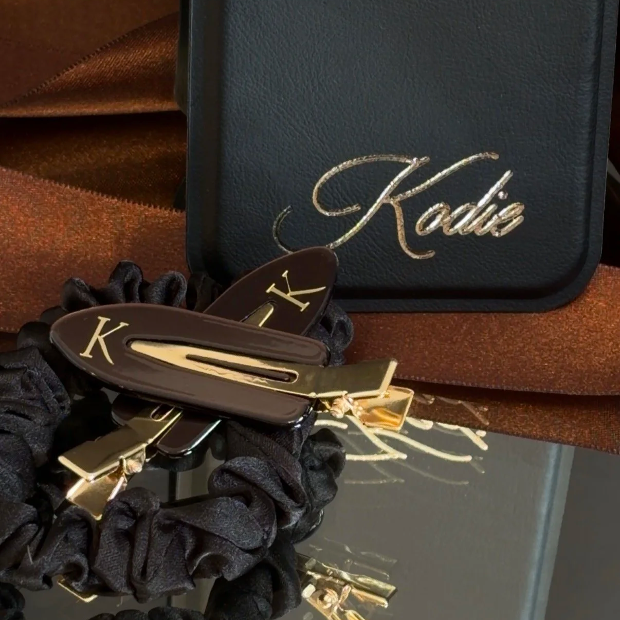 A black case with the name 'Kodie' in gold cursive embroidery, a hair scrunchie, and decorative hair clips with a gold and black design, all placed on a metallic surface.