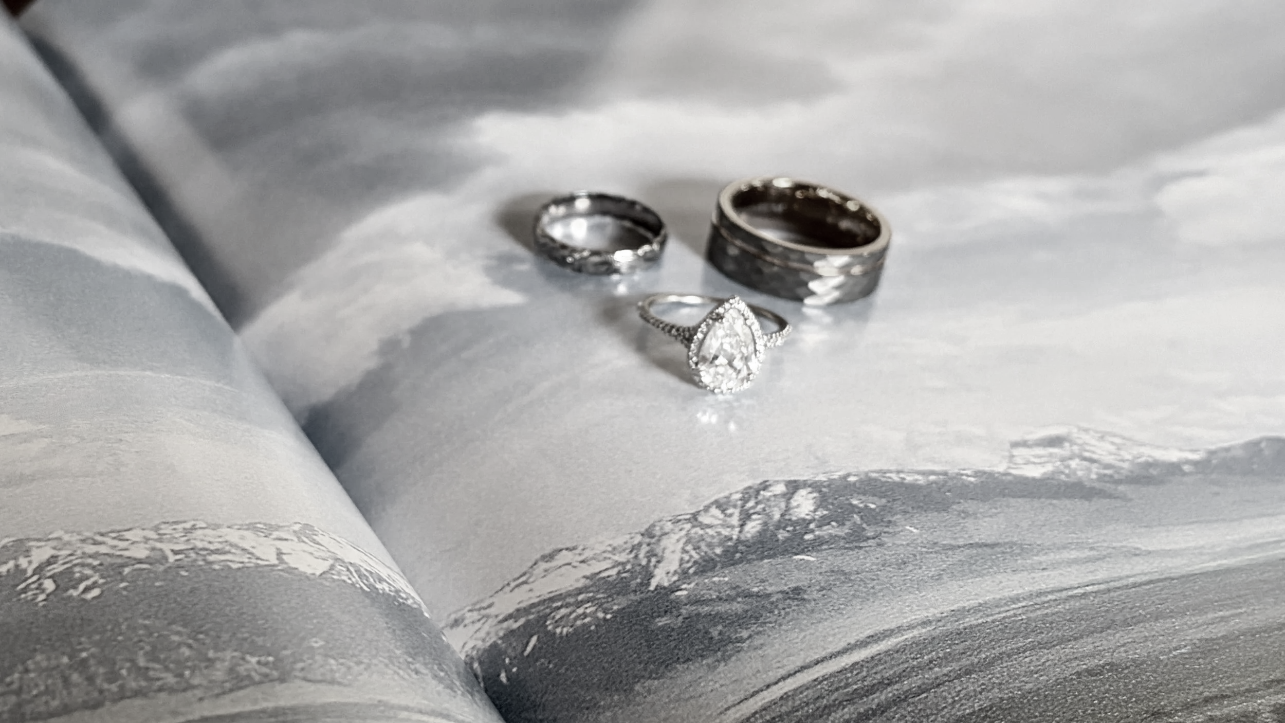 Three silver rings, one with a large teardrop-shaped gemstone surrounded by smaller stones, resting on an open magazine or book with black and white pages.