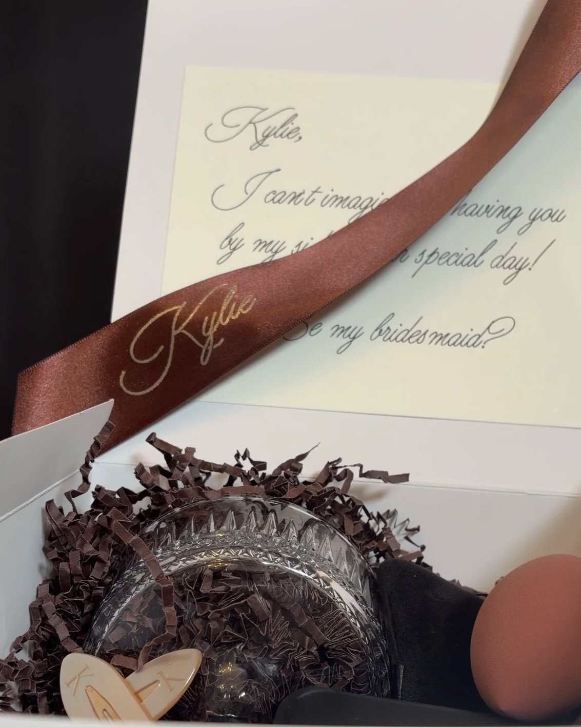 Close-up of a wedding gift box containing a clear glass item, tissue paper, and a brown ribbon with the name 'Kylie' written in gold, along with a heartfelt note.