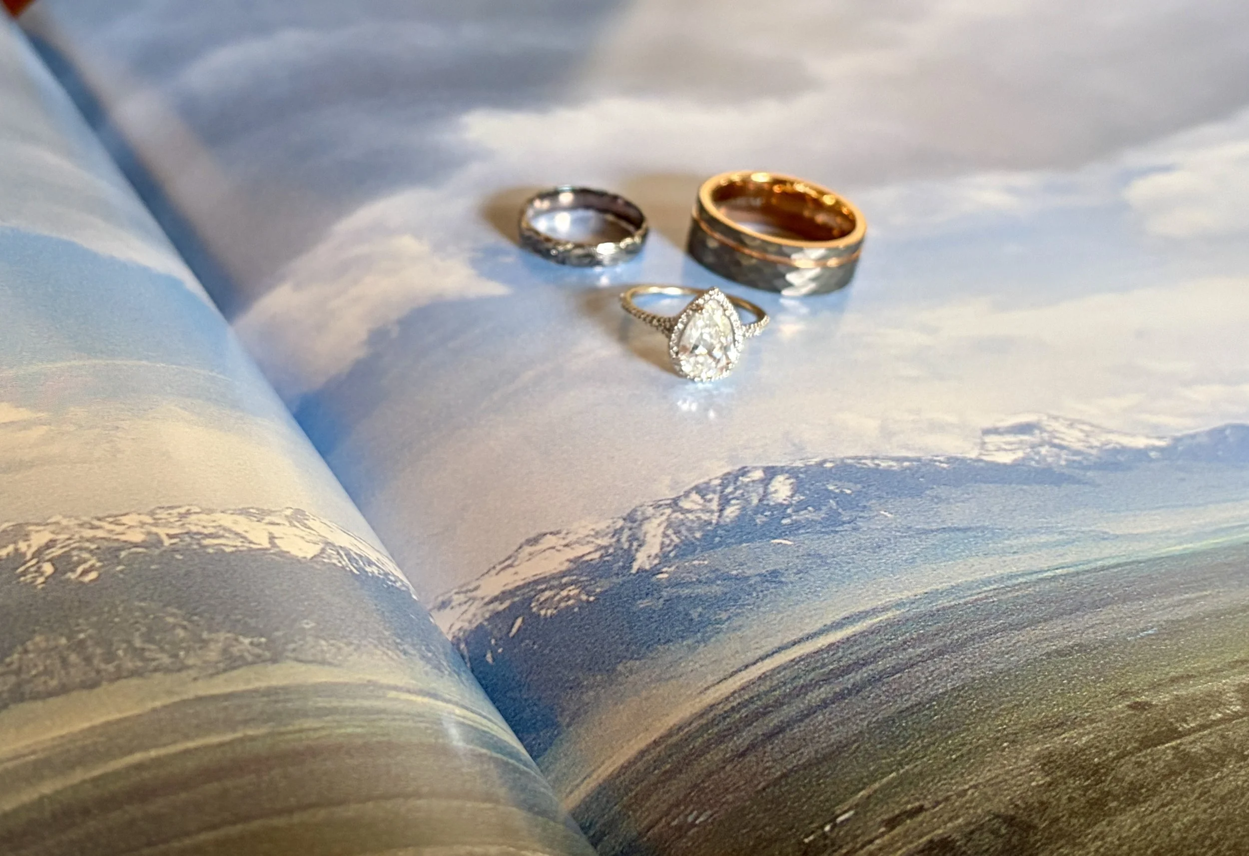 Three rings, one with a large teardrop-shaped diamond surrounded by smaller diamonds, and two plain bands, are resting on a surface resembling an open book with mountain and sky imagery.