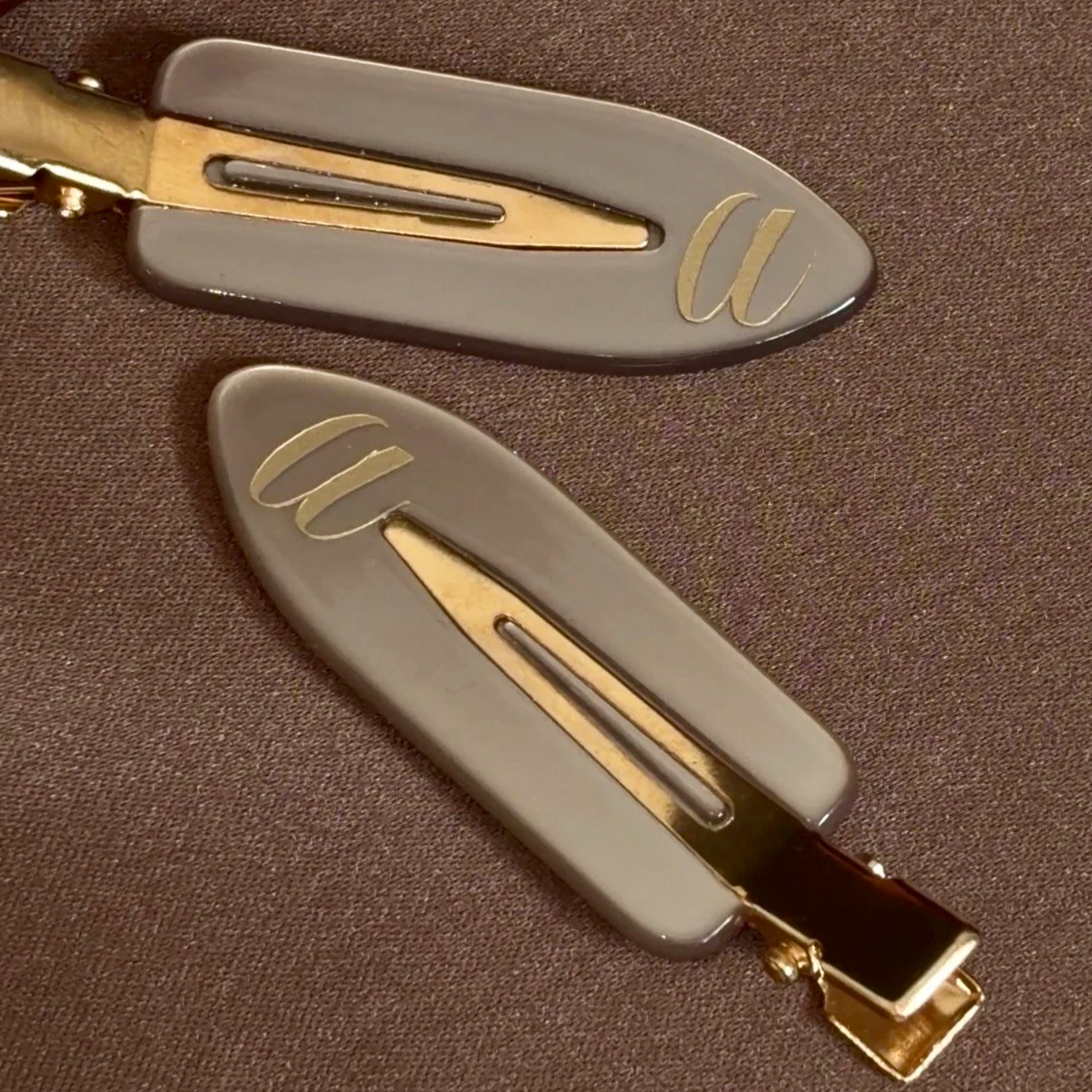 Two hairclips with gray shells and gold accents placed on a brown fabric surface.