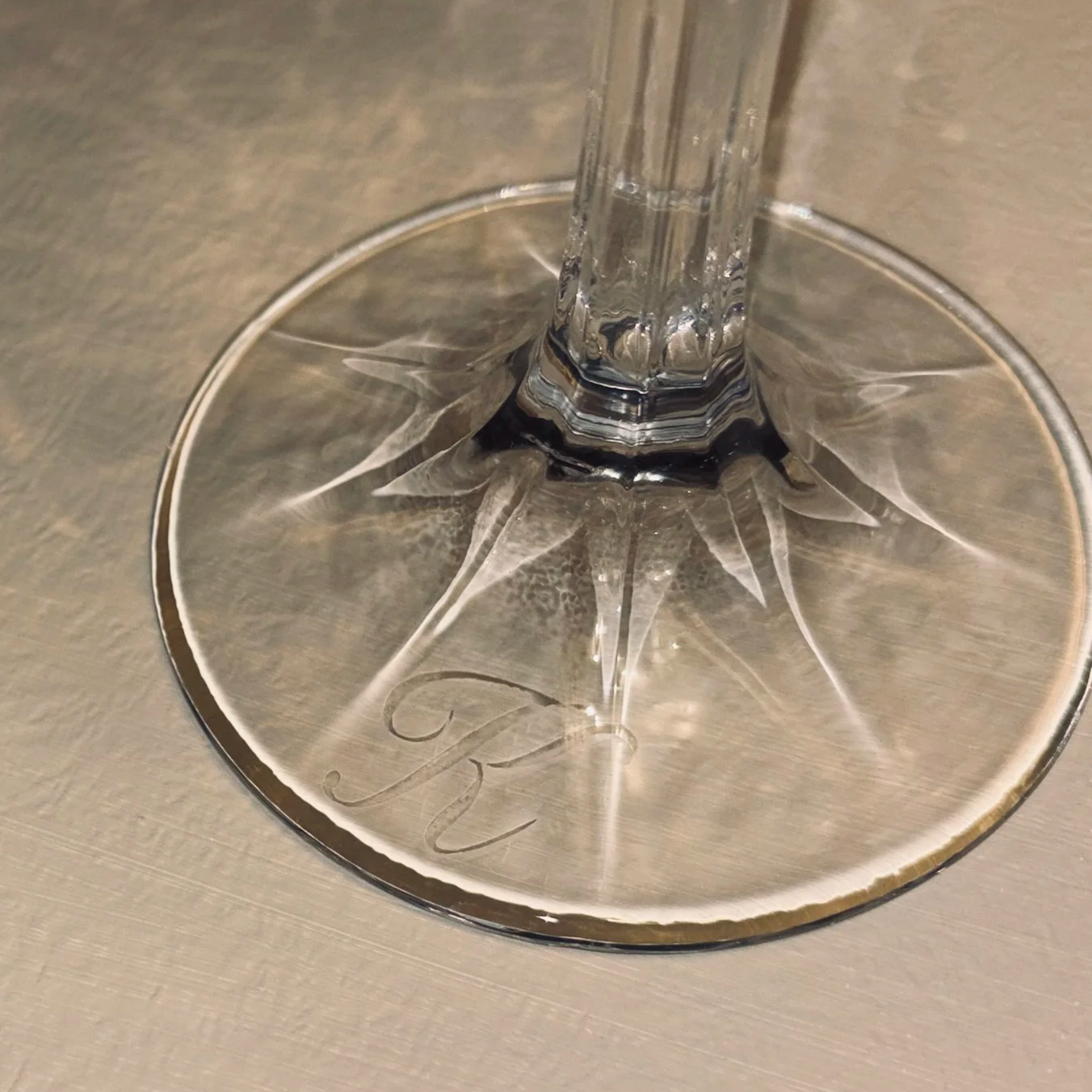 Close-up of a glass champagne flute with a long stem, resting on a round glass coaster with an engraved letter. The glass base has an etched 'K' on it. The surface appears to be a metallic or reflective table.