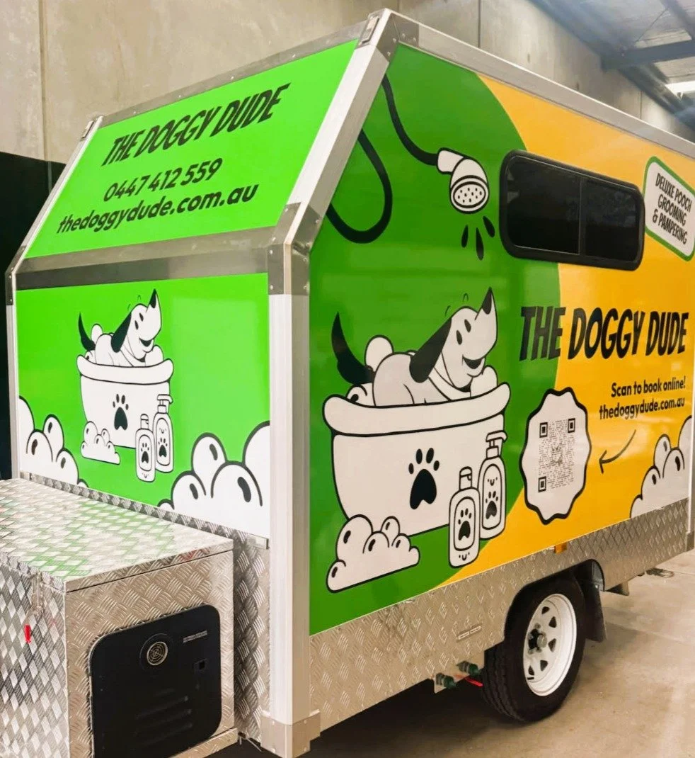 A colorful food truck called 'The Doggy Dude' with cartoon graphics of a dog in a bathtub with shampoo bottles, and paw prints. The truck has a green and yellow design with a website and phone number. It offers grooming and pet care services, with a QR code for booking.