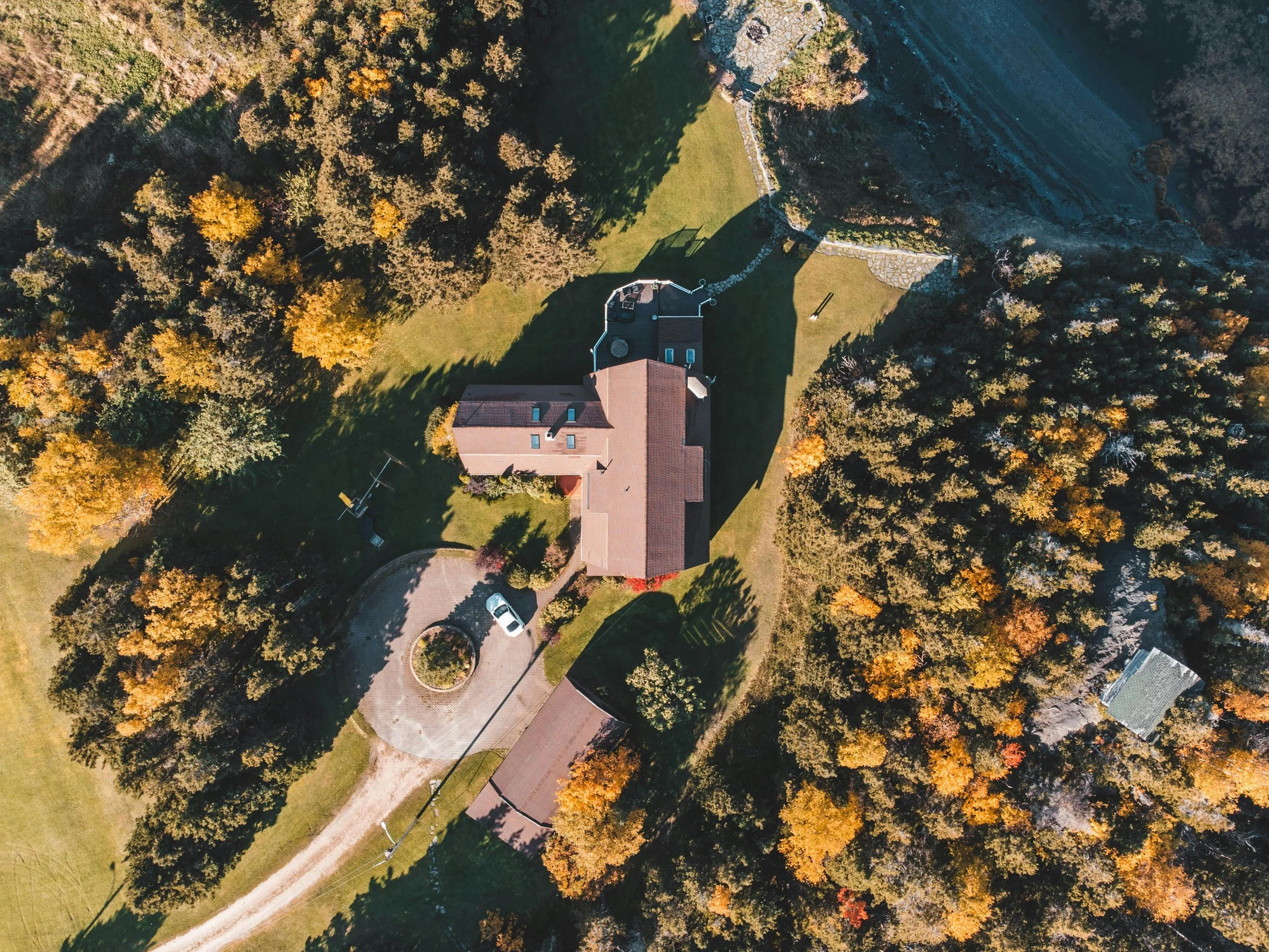 An aerial view of a residential property surrounded by autumn-colored trees, with a curved driveway, a lawn, and a house with a red roof.
