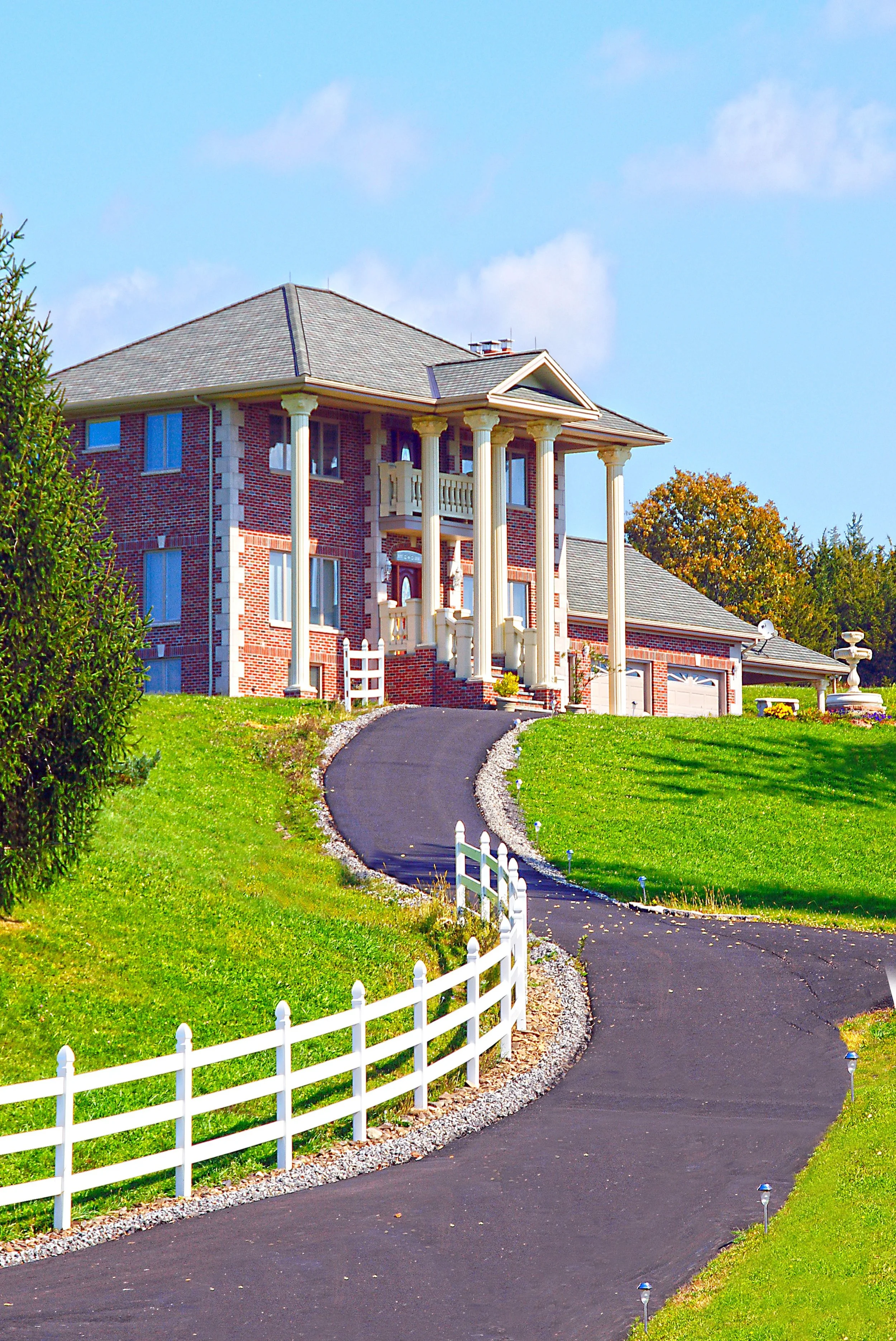 A large brick house with white columns, a grey roof, and a driveway leading up to it. There are green lawns, trees, and a white picket fence along the driveway. The sky is blue with some clouds.