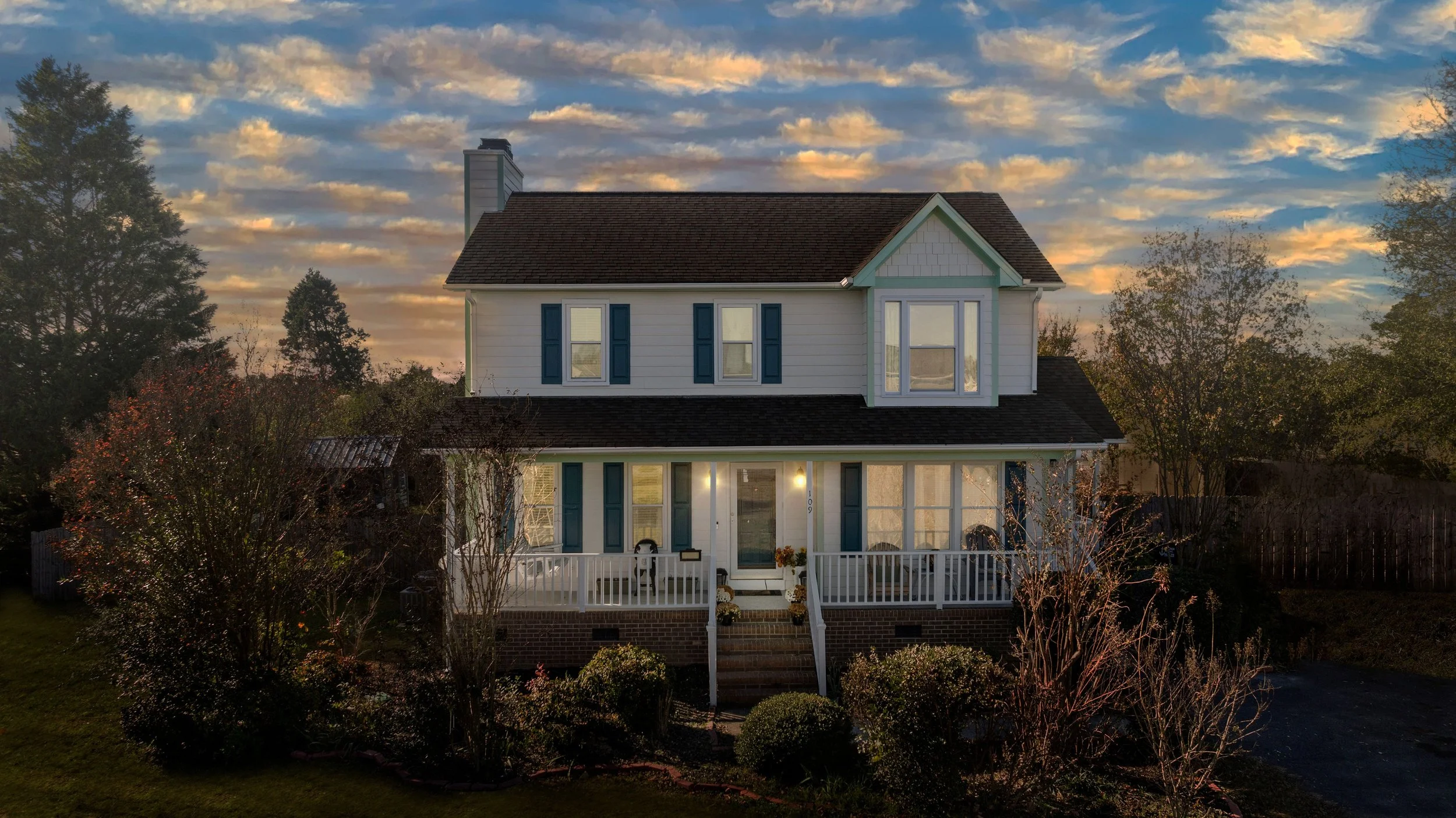 A two-story house with a front porch, white siding, blue shutters, and a gabled roof, surrounded by trees and a landscaped yard at sunset.