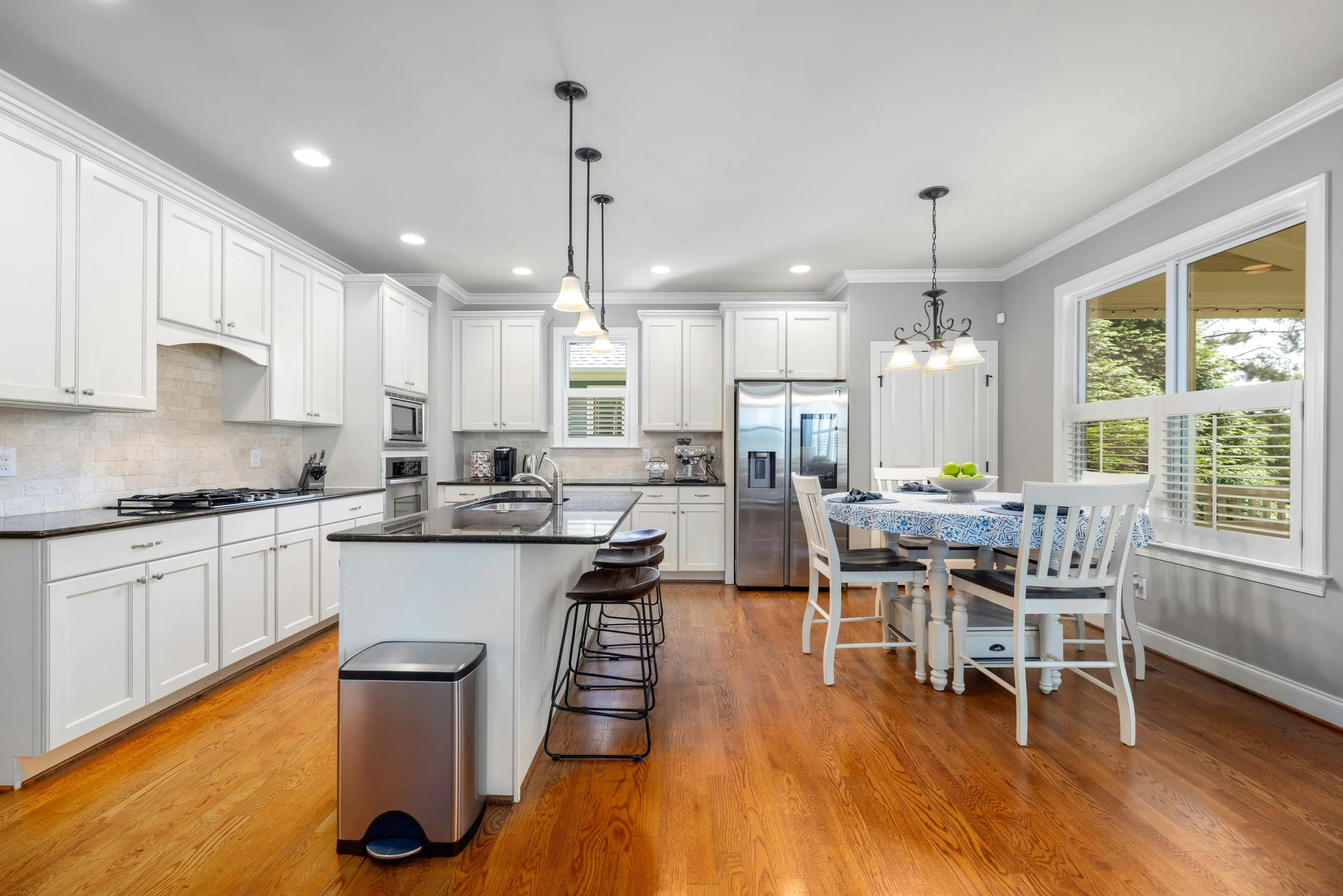 Bright kitchen with white cabinets, stainless steel refrigerator, island with bar stools, and a dining table with chairs near large windows with white shutters.