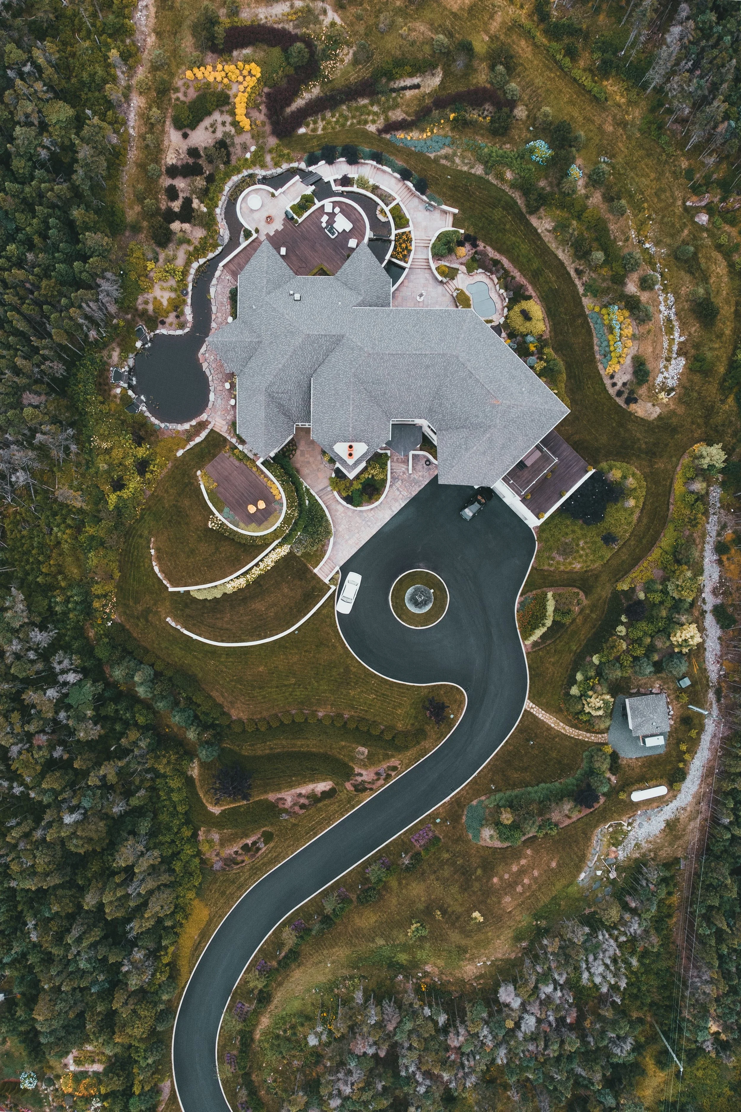An aerial view of a large modern house with a dark roof, surrounded by landscaped gardens, a small pond, and a winding driveway in a lush, wooded area.