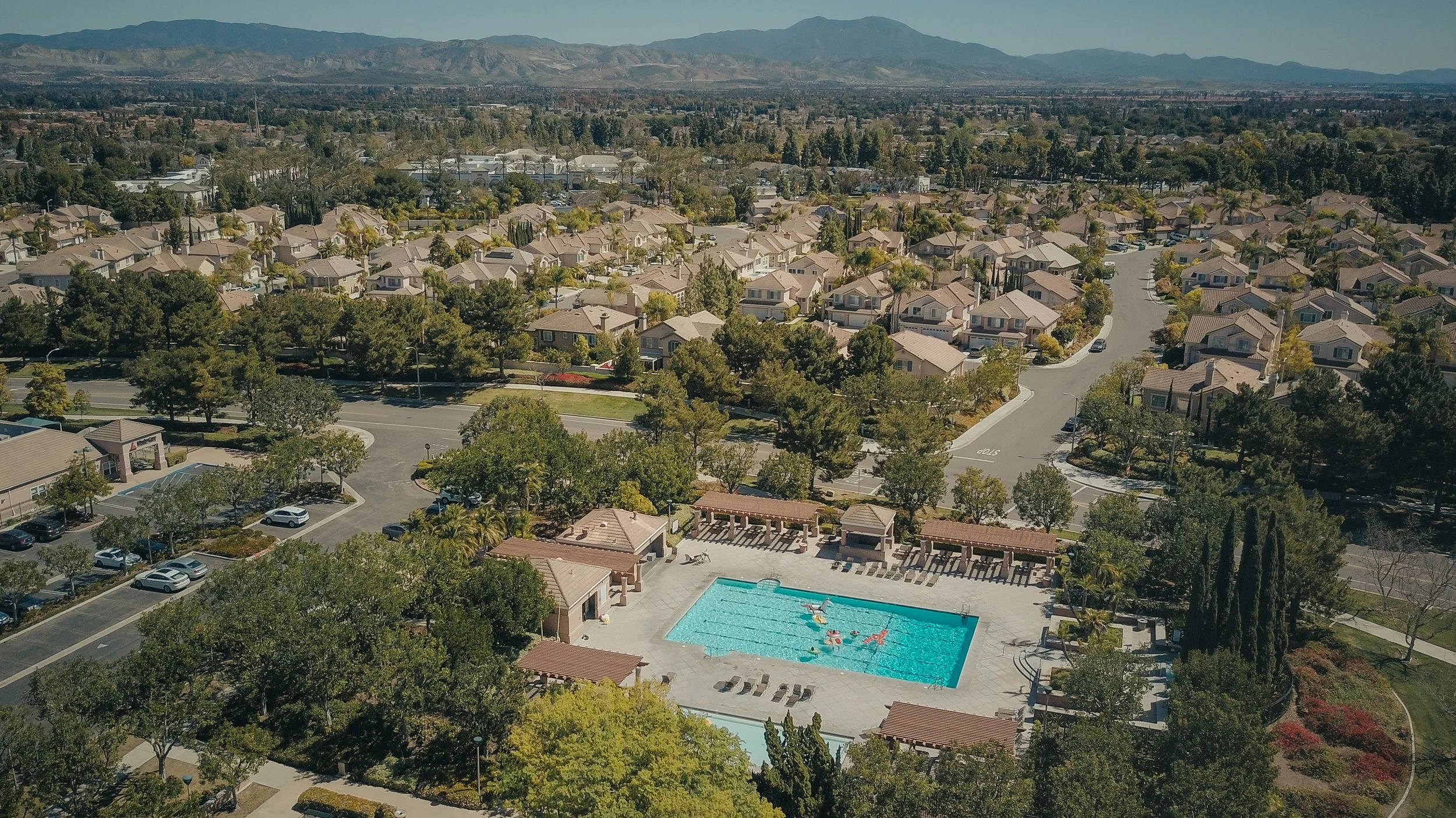 Aerial view of a residential community with single-family homes, a swimming pool, parking lots, and surrounding trees, with mountains in the background.