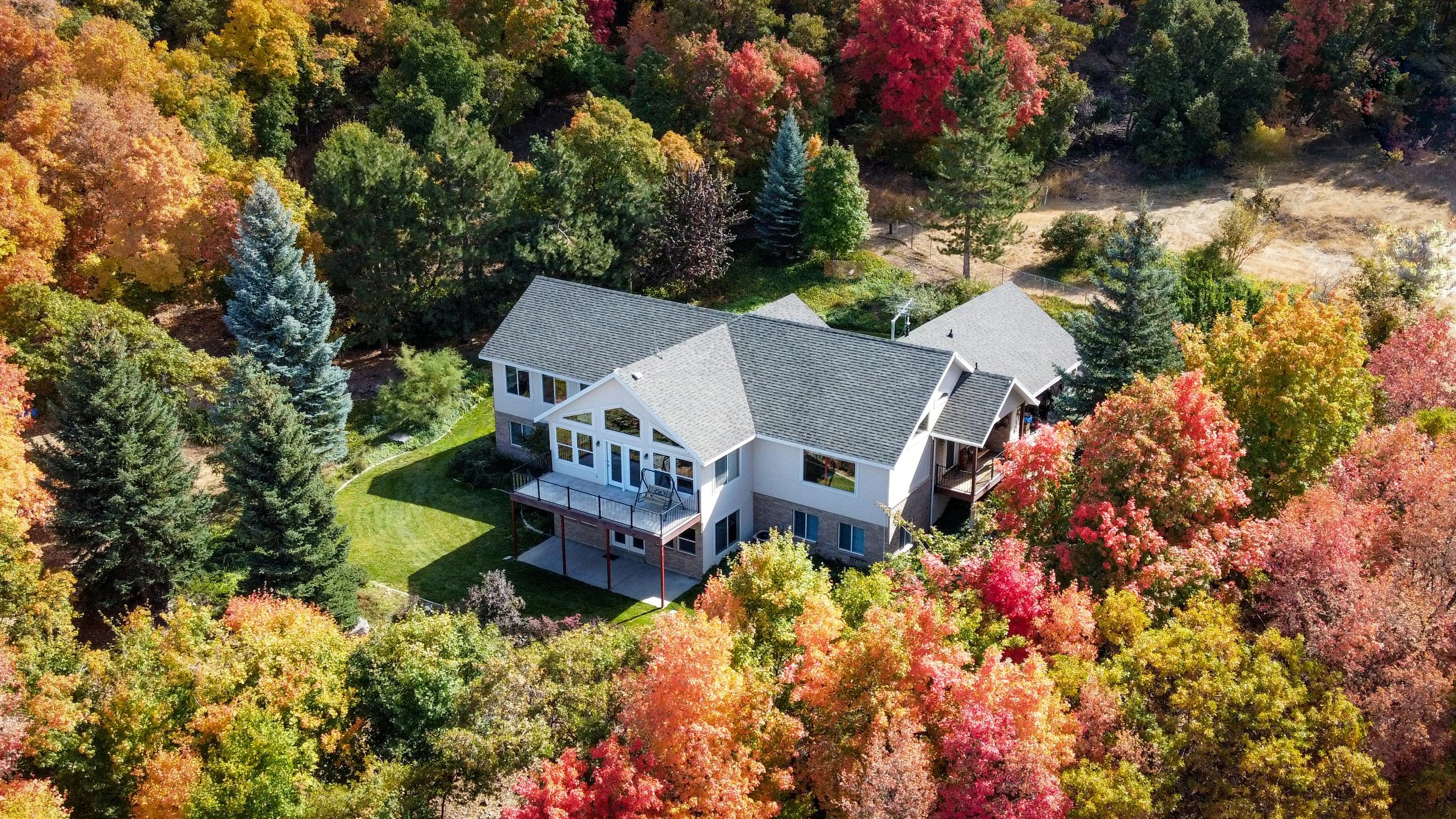 An aerial view of a modern house surrounded by colorful fall foliage with green, red, orange, and yellow leaves, and a grassy yard.