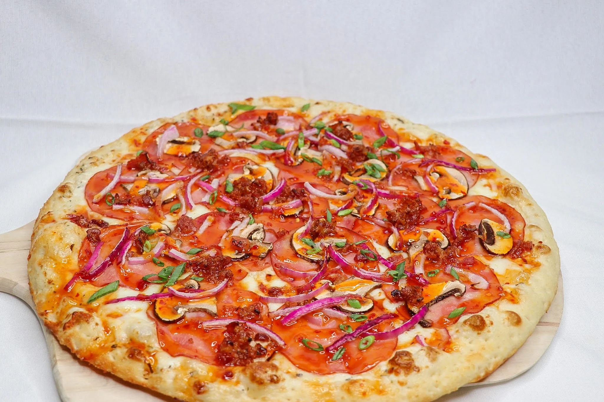 A whole pizza topped with sliced tomatoes, red onions, ground meat, black olives, and chopped green herbs on a wooden pizza peel.