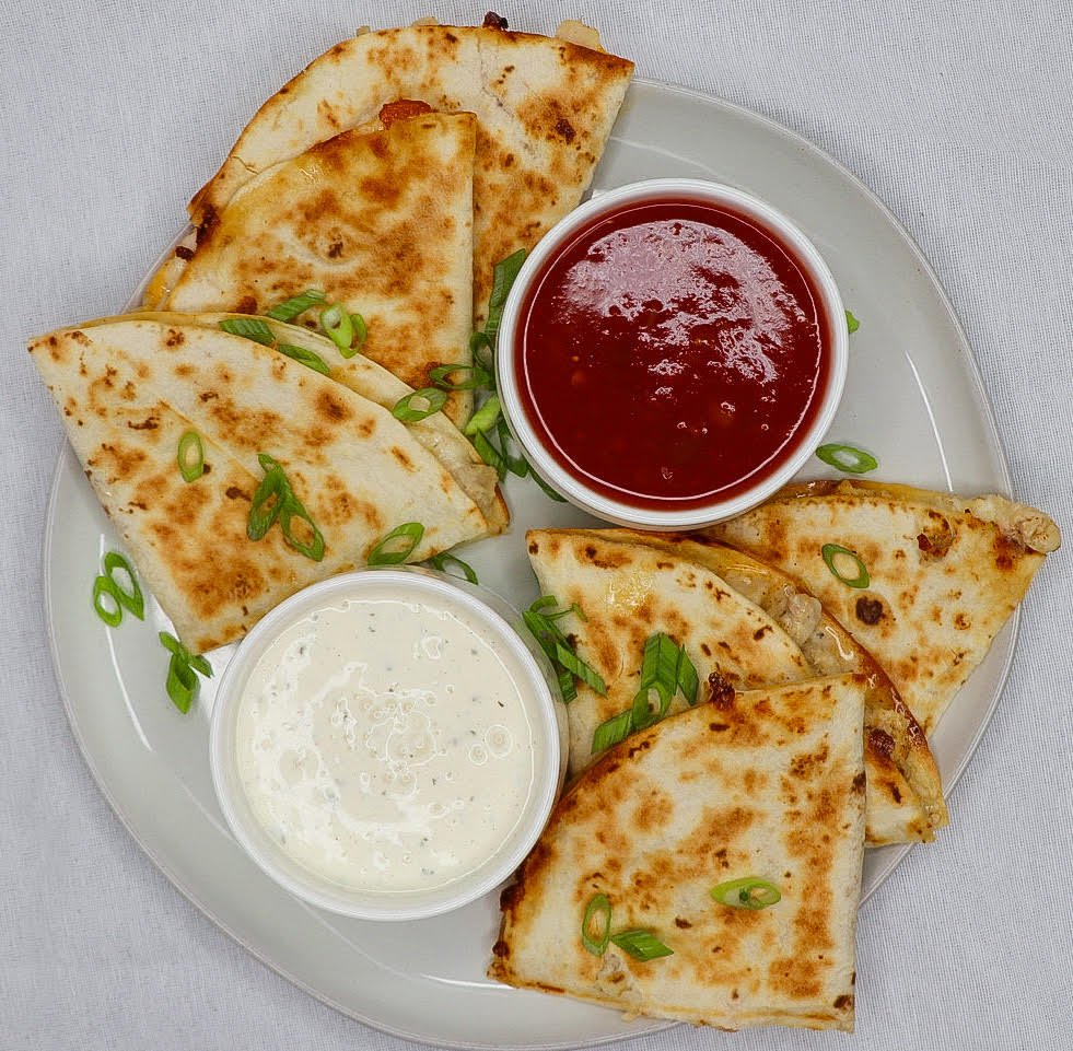 Sliced quesadillas garnished with chopped green onions, served with red salsa and sour cream on a white plate.