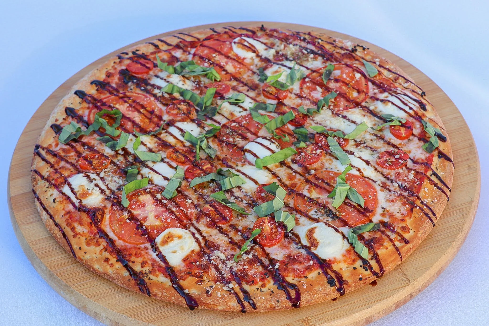 A pizza with tomatoes, fresh basil, mozzarella, drizzled with balsamic glaze, on a round wooden board.