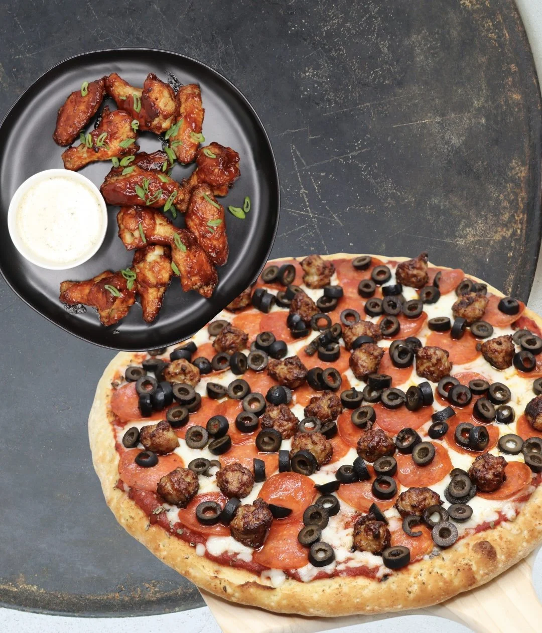 A pizza topped with pepperoni, black olives, and sausage, along with a side of buffalo chicken wings with green onions and a small cup of dipping sauce.