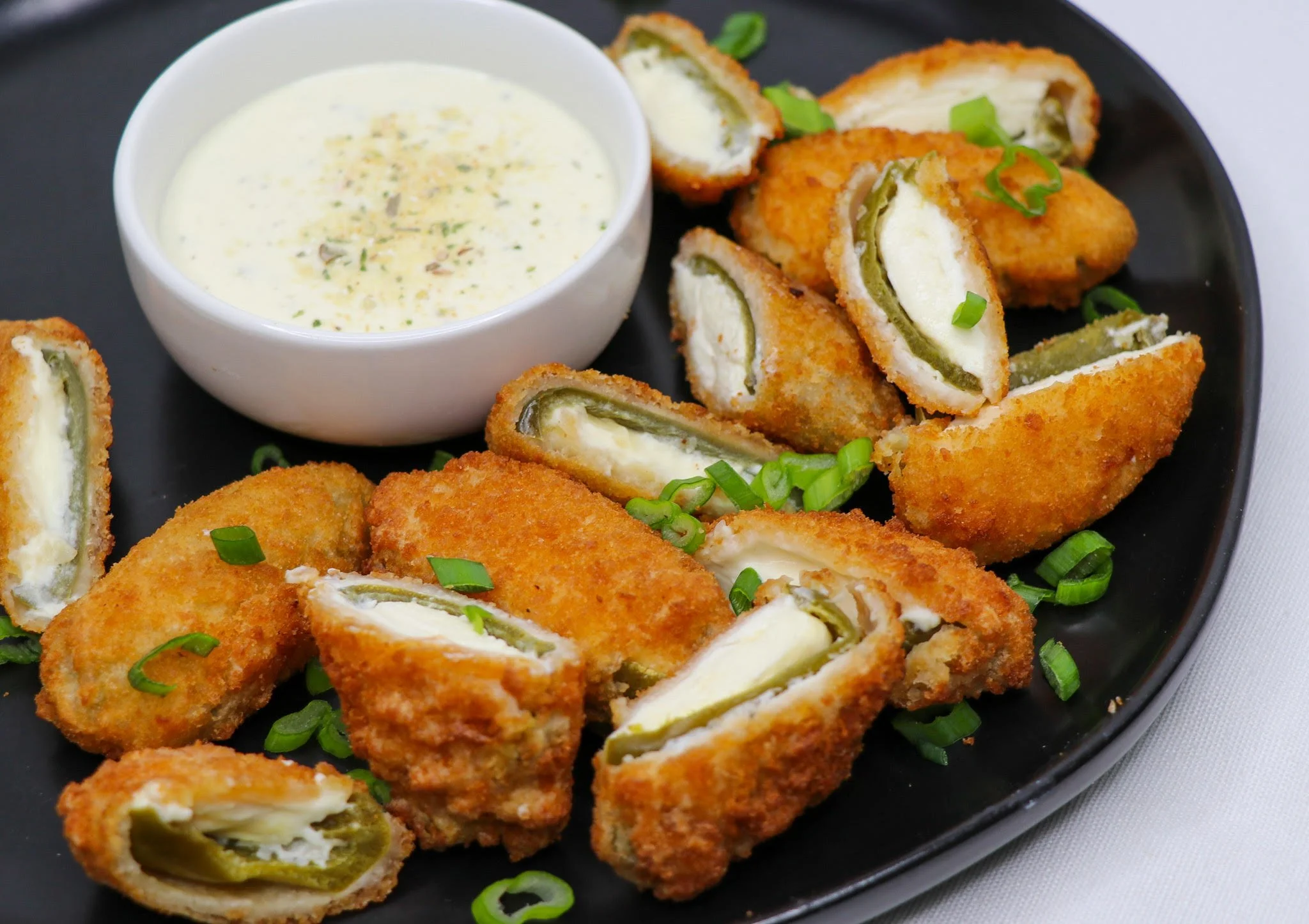 Fried jalapeño poppers with a side of dipping sauce on a black plate, garnished with chopped green onions.