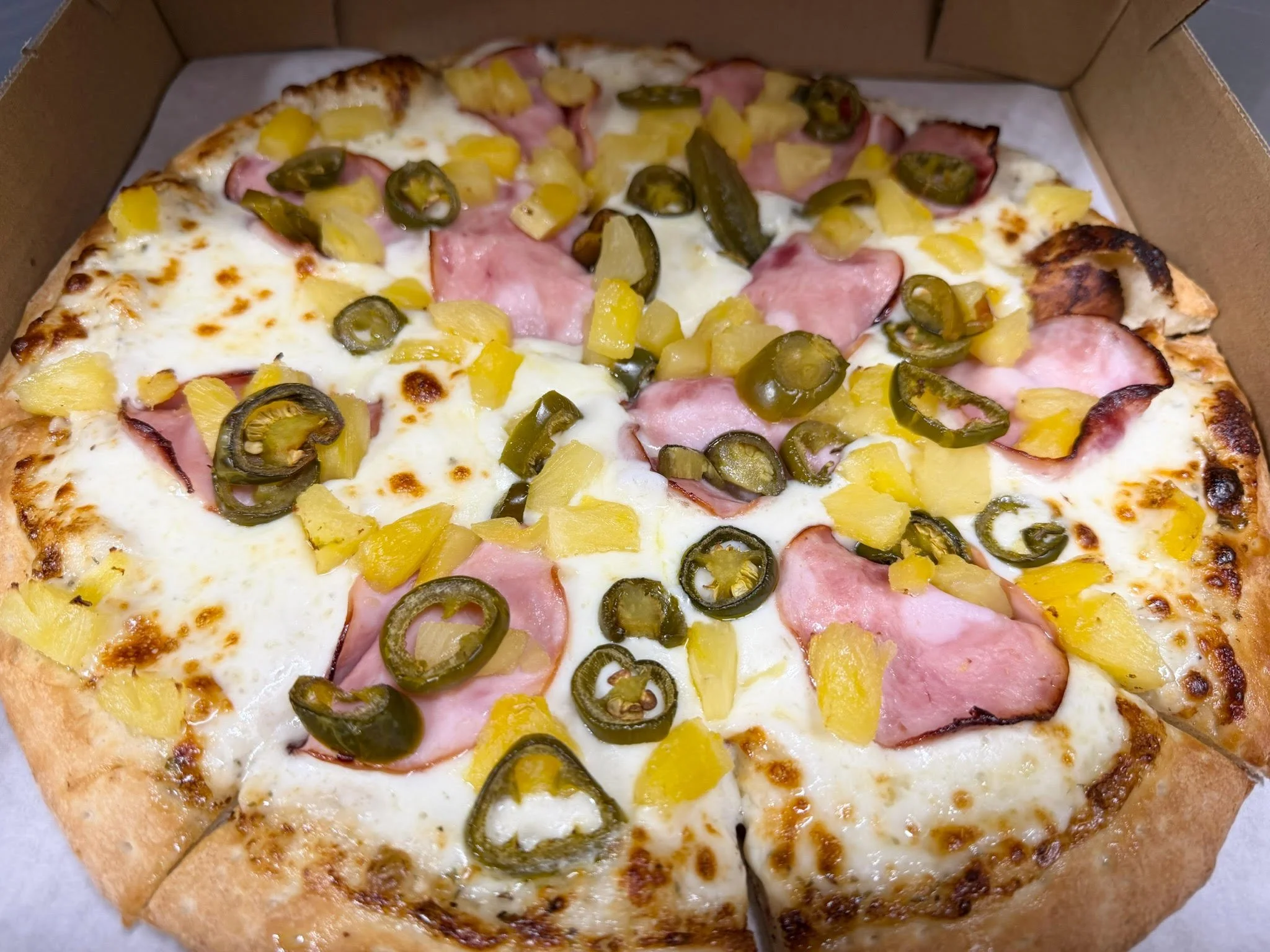 A pizza with cheese, pepperoni, pineapple chunks, and sliced green jalapeño peppers in a cardboard box.