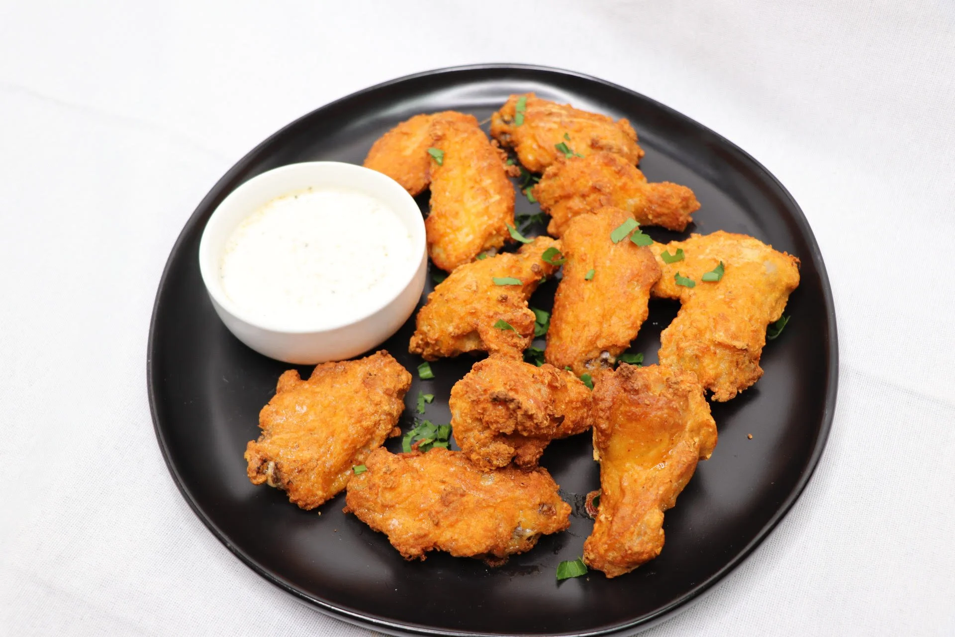Fried chicken wings on a black plate with chopped green herbs and a side of creamy dipping sauce.