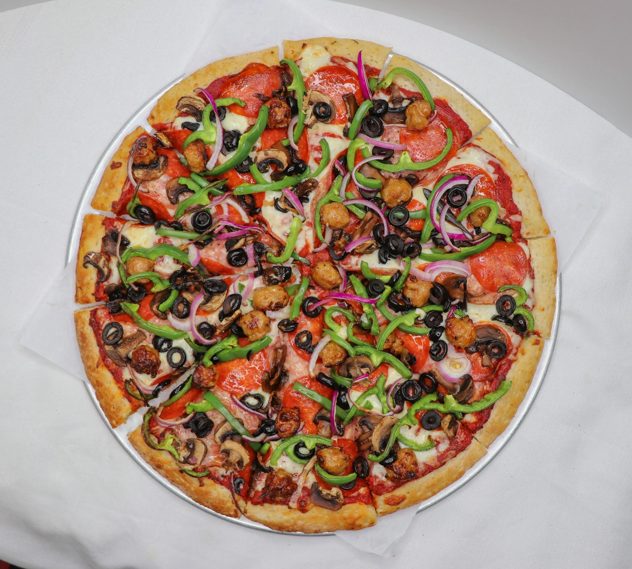 A pizza with pepperoni slices, sliced green bell peppers, onions, black olives, mushrooms, and chunks of sausage on a pizza crust with melted cheese, served on a round metal tray.