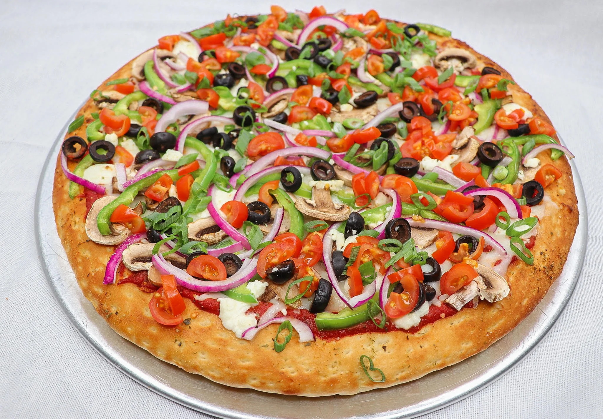 A pizza topped with cherry tomatoes, sliced green bell peppers, sliced red onions, black olives, sliced mushrooms, and chopped green onions on a thin crust. The pizza is on a silver tray.