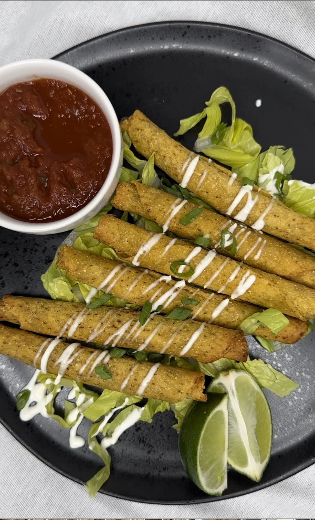 Fried fish strips garnished with chopped green onions, drizzled with white sauce, served on a bed of lettuce with lime wedges and a side of red salsa.