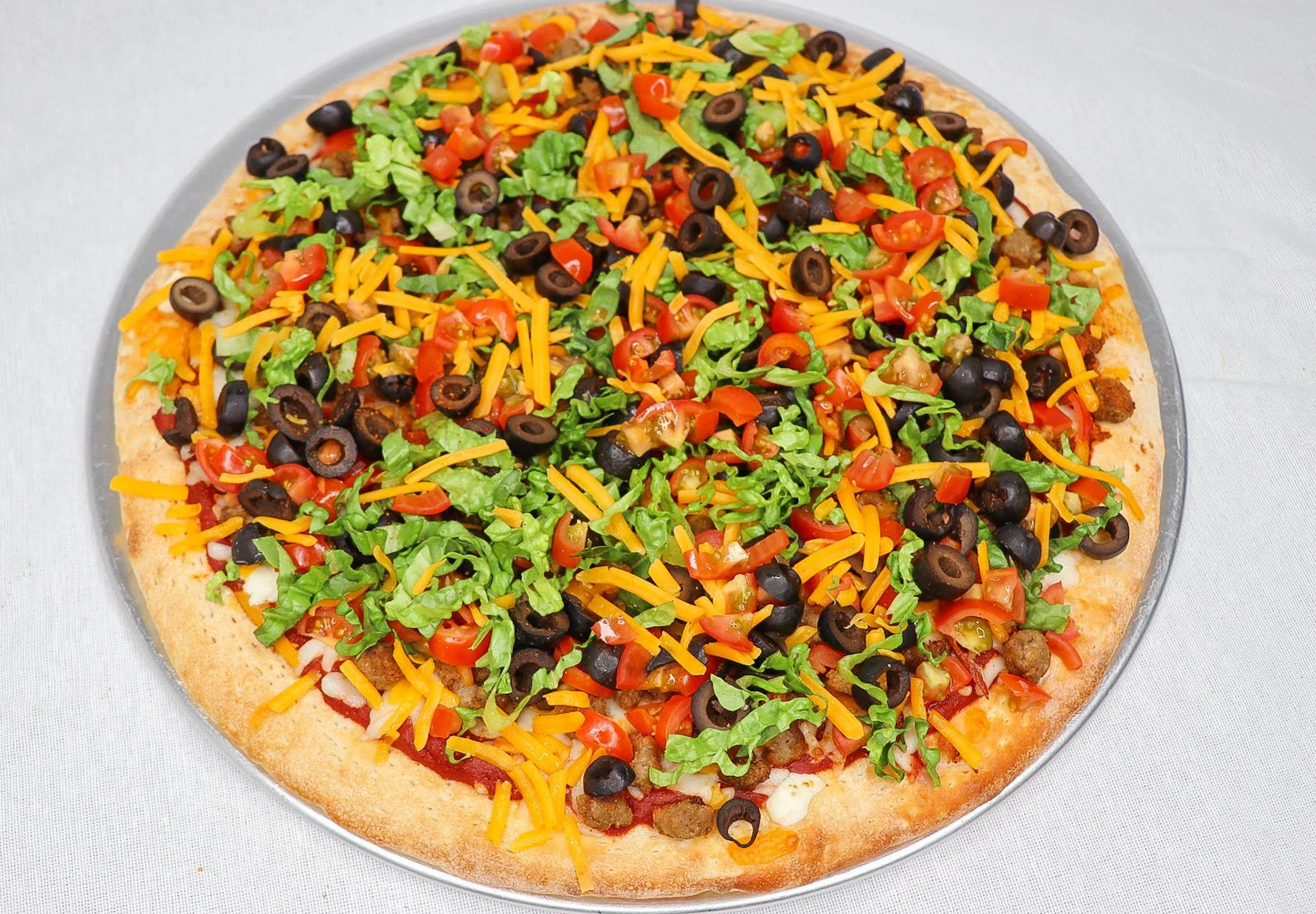 A pizza topped with shredded cheddar cheese, chopped red tomatoes, black olives, green lettuce, and cooked ground beef on a crust, on a round silver tray.
