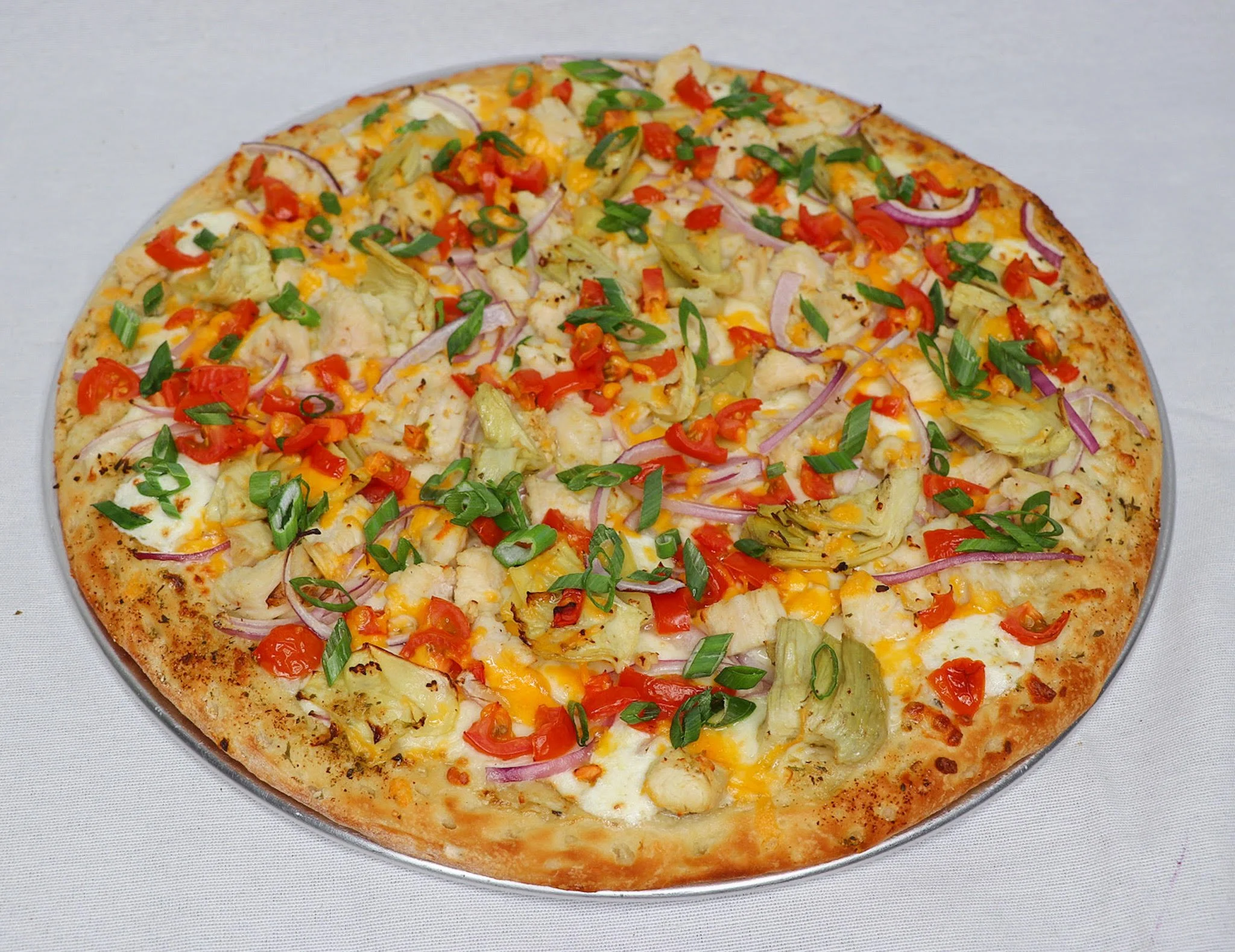 A pizza topped with chopped red onions, red bell peppers, chopped green onions, pineapple pieces, shredded cheese, and chicken, on a metallic pizza tray.