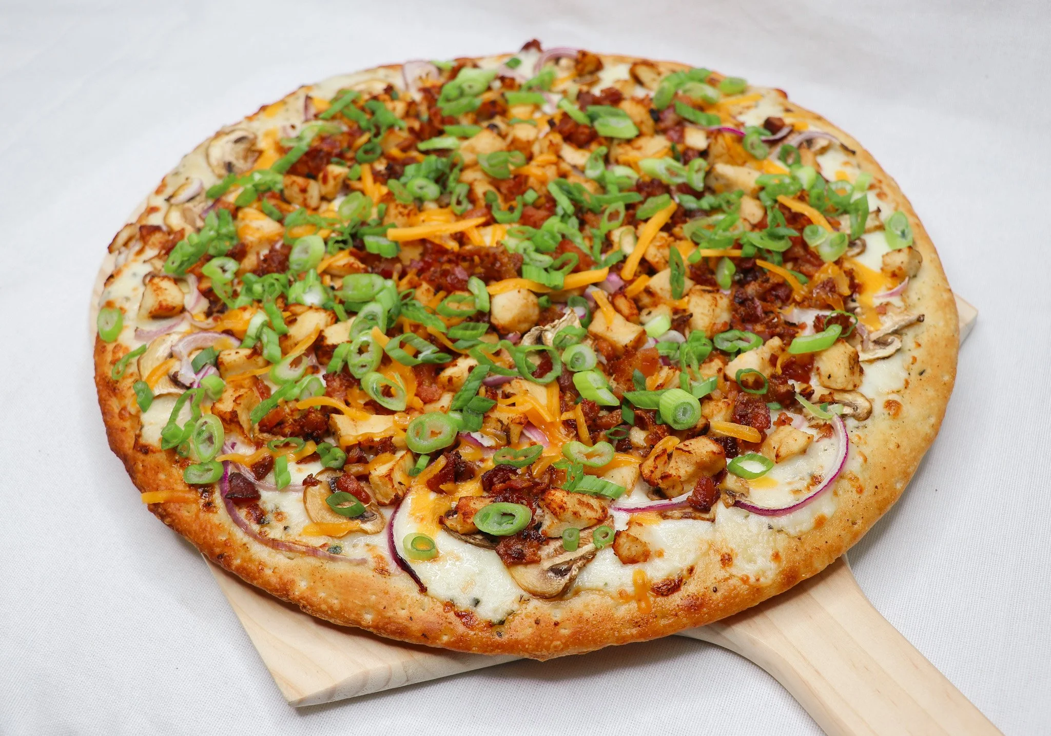 A freshly baked pizza with cheese, pepperoni, cooked chicken pieces, sliced red onions, chopped green onions, and mushrooms on a white surface.
