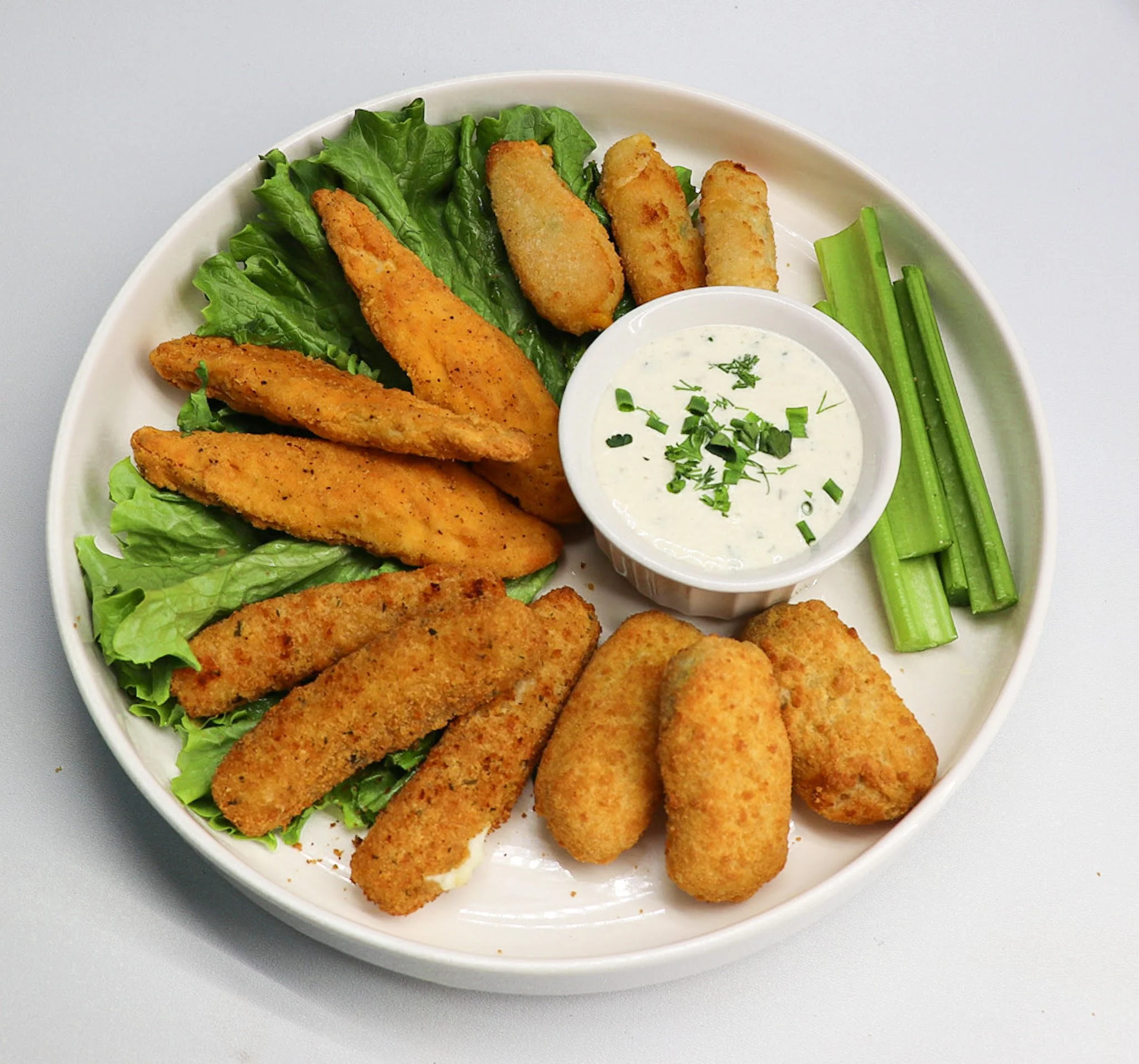 White oval plate with fried fish fillets, celery sticks, and a small bowl of creamy dipping sauce garnished with chopped herbs and green onions on a gray surface.