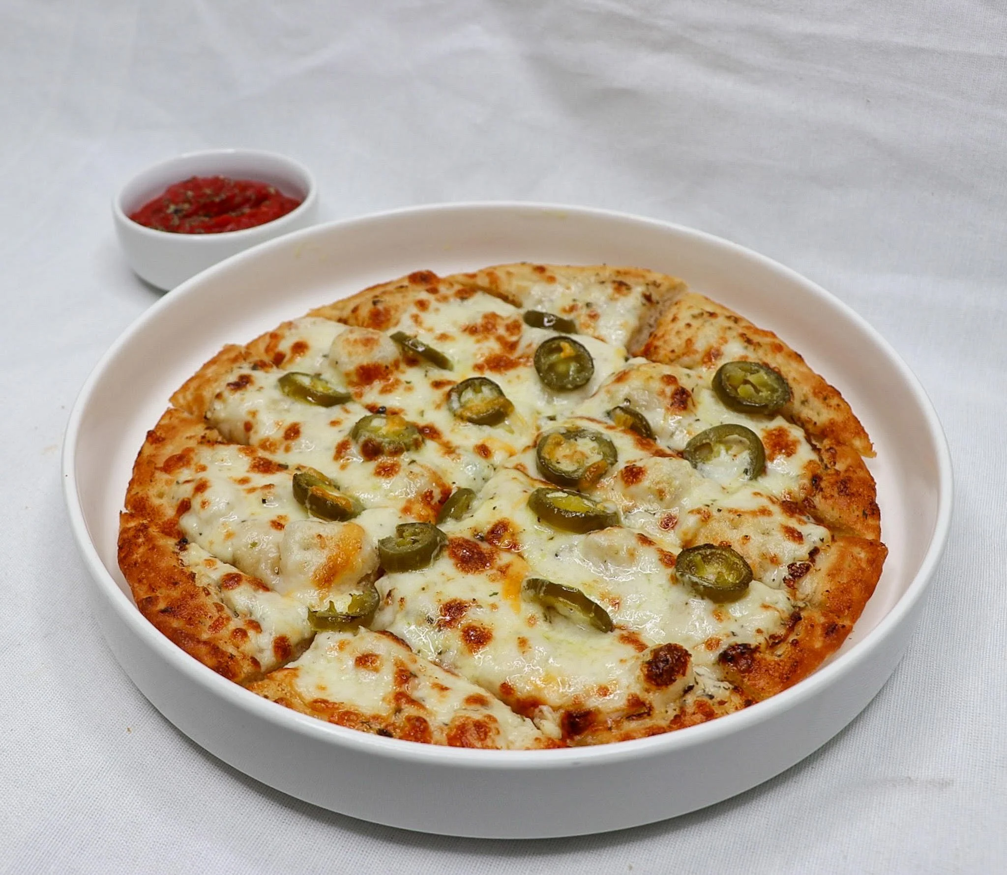A cheese pizza topped with sliced green jalapeño peppers on a white plate, with a small bowl of red sauce in the background on a white surface.
