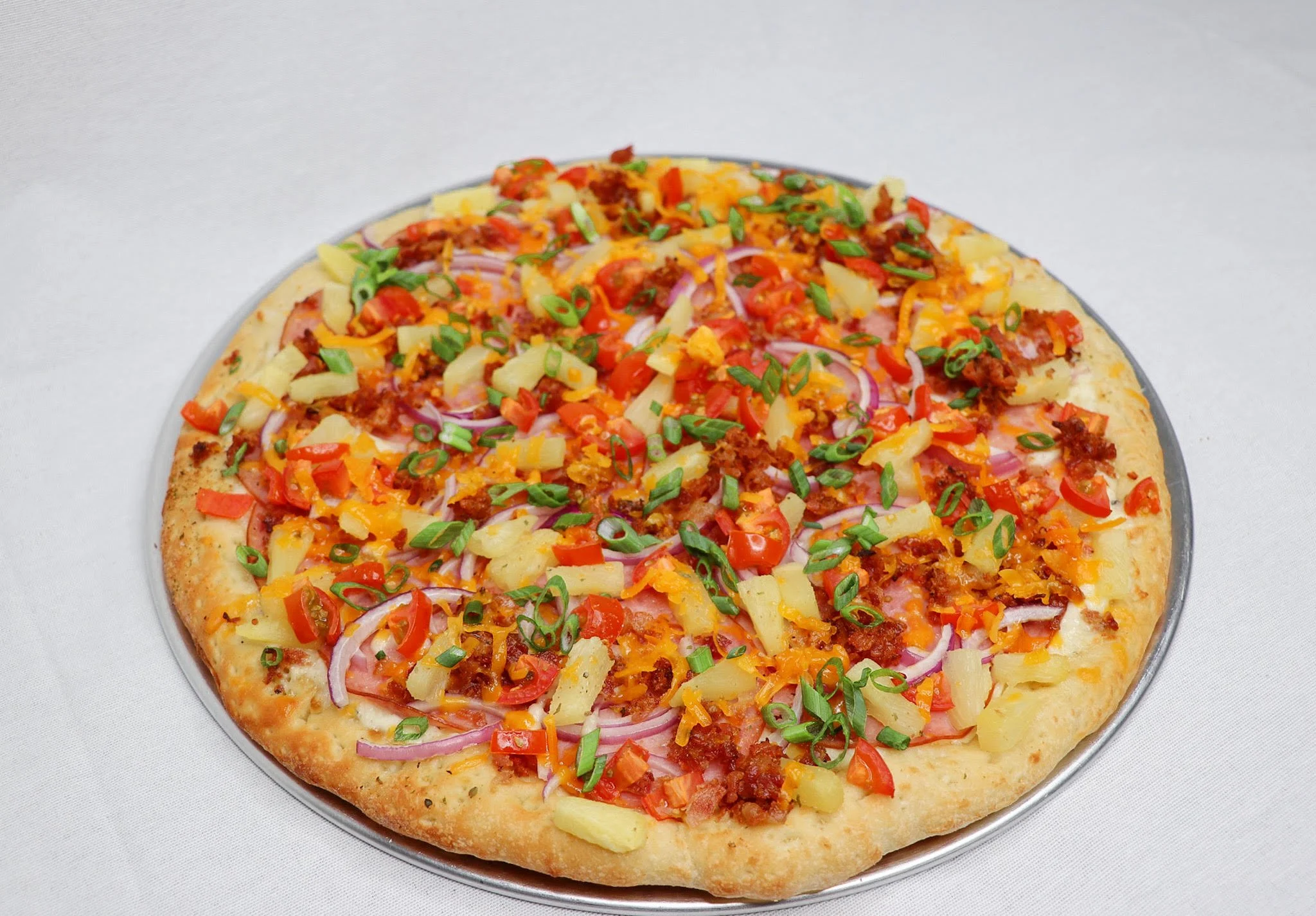 A pizza with various toppings, including cheese, pineapple, red onions, chopped tomatoes, green onions, and ground meat, on a round tray against a white background.