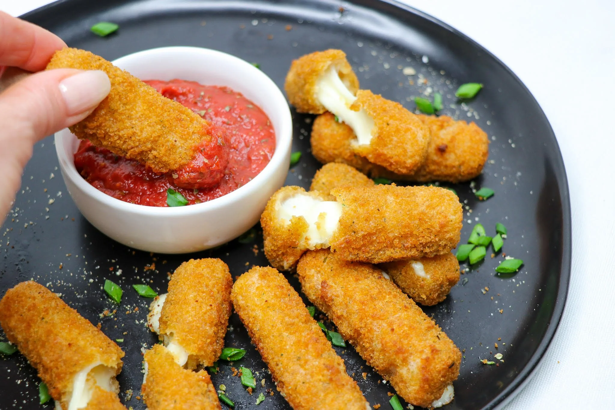 Breaded mozzarella sticks with some cut in half displaying melted cheese inside, served with marinara sauce for dipping, garnished with chopped green onions on a black plate.