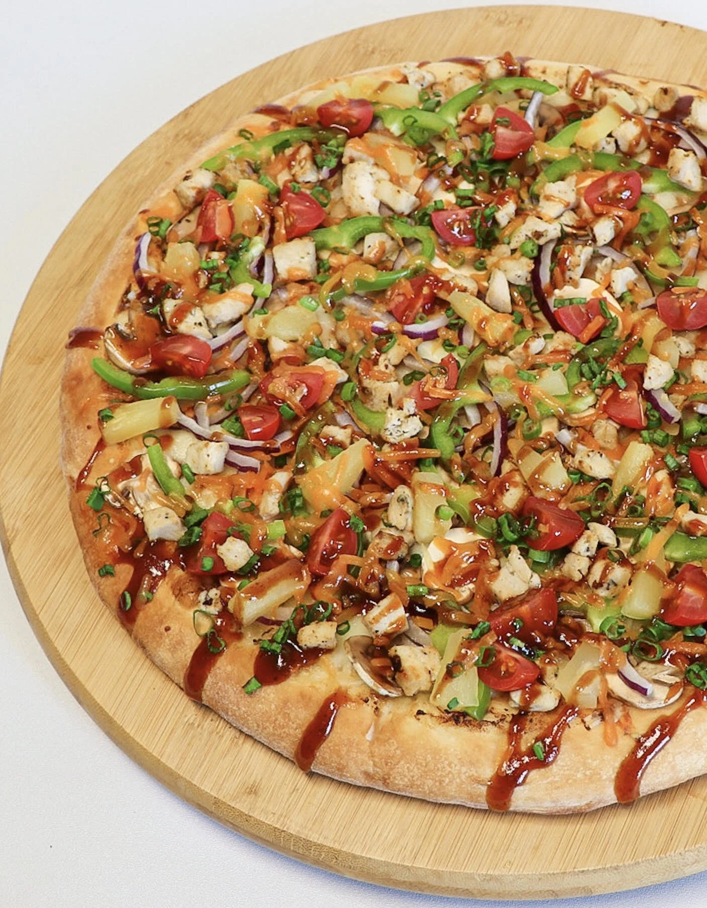 A pizza topped with chopped grilled chicken, cherry tomatoes, green bell peppers, pineapple, red onions, chopped green onions, and drizzled with barbecue sauce, on a round wooden pizza board.