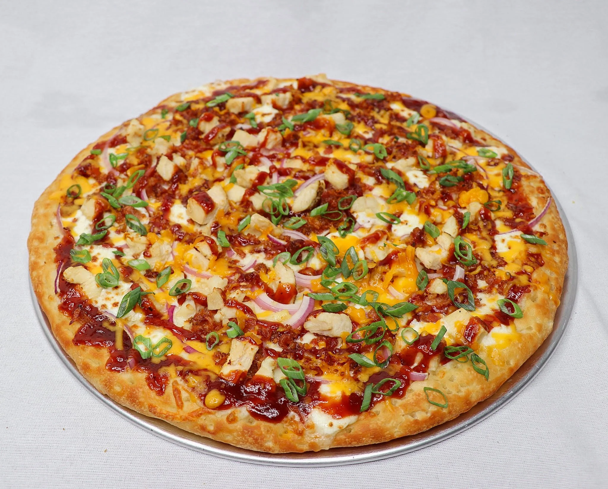 A pizza topped with chopped green onions, diced chicken, shredded cheese, bacon bits, and drizzled with barbecue sauce, served on a metal tray.