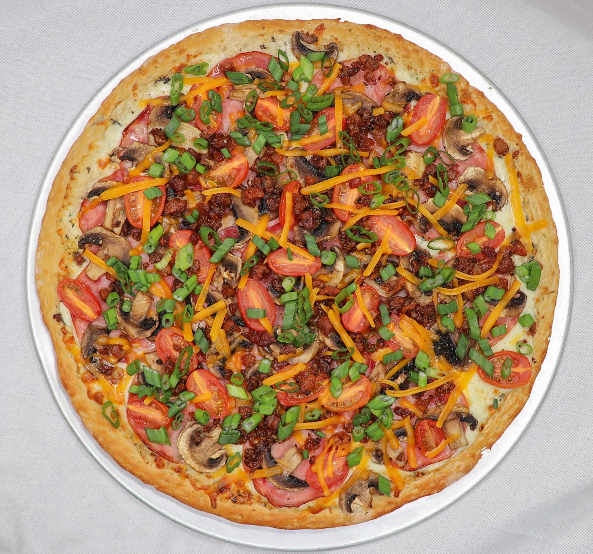 A pizza with a golden crust topped with cherry tomatoes, sliced mushrooms, shredded cheese, chopped green onions, chopped bacon, and ground meat, served on a silver tray.