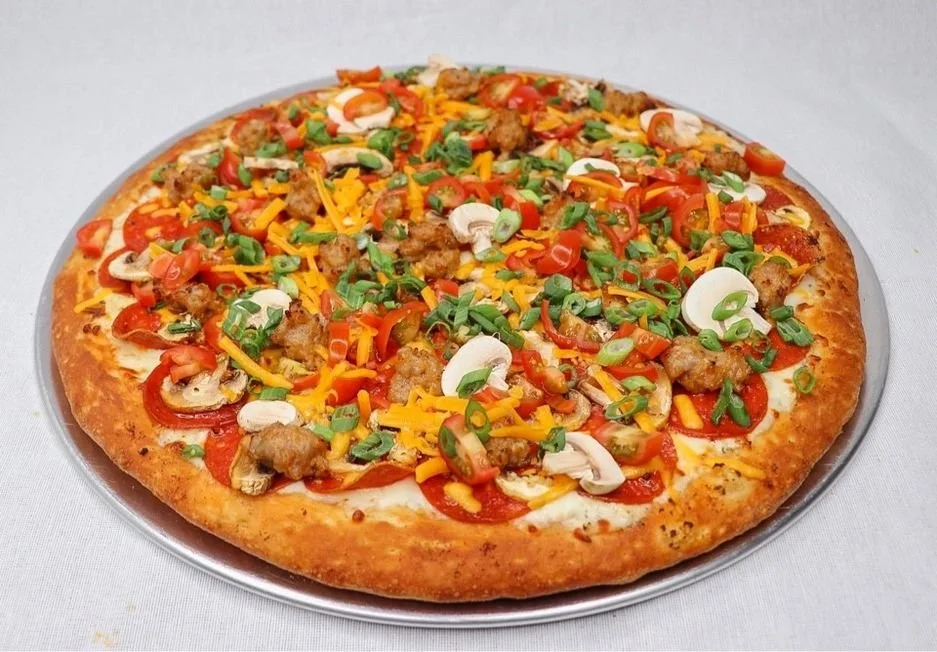 A freshly baked pizza topped with sausage, red and yellow peppers, mushrooms, chopped green onions, and shredded cheddar cheese on a round metal tray.