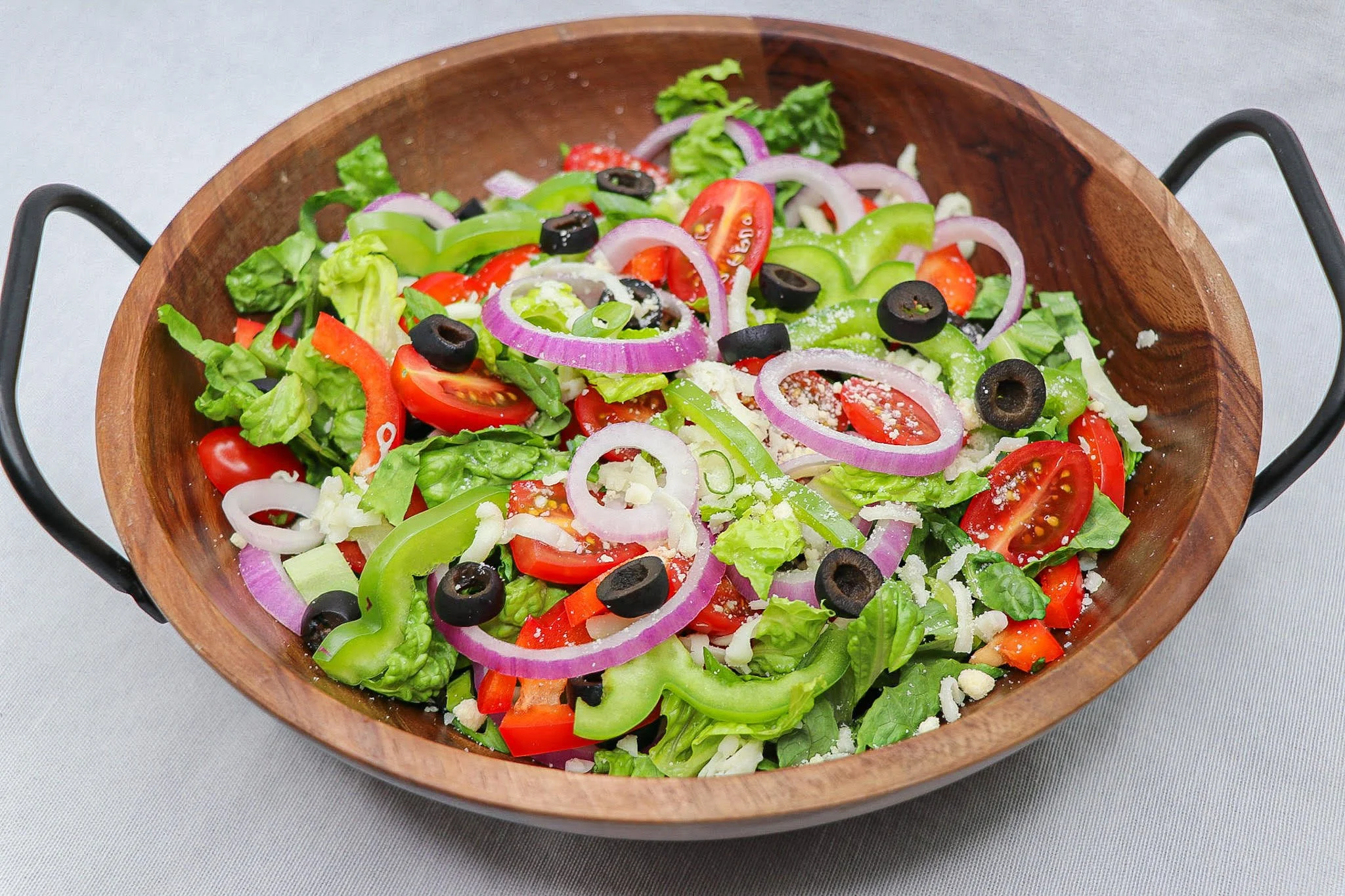A wooden bowl filled with fresh salad containing cherry tomatoes, sliced red onions, black olives, green bell peppers, and shredded cheese on a light grey surface.
