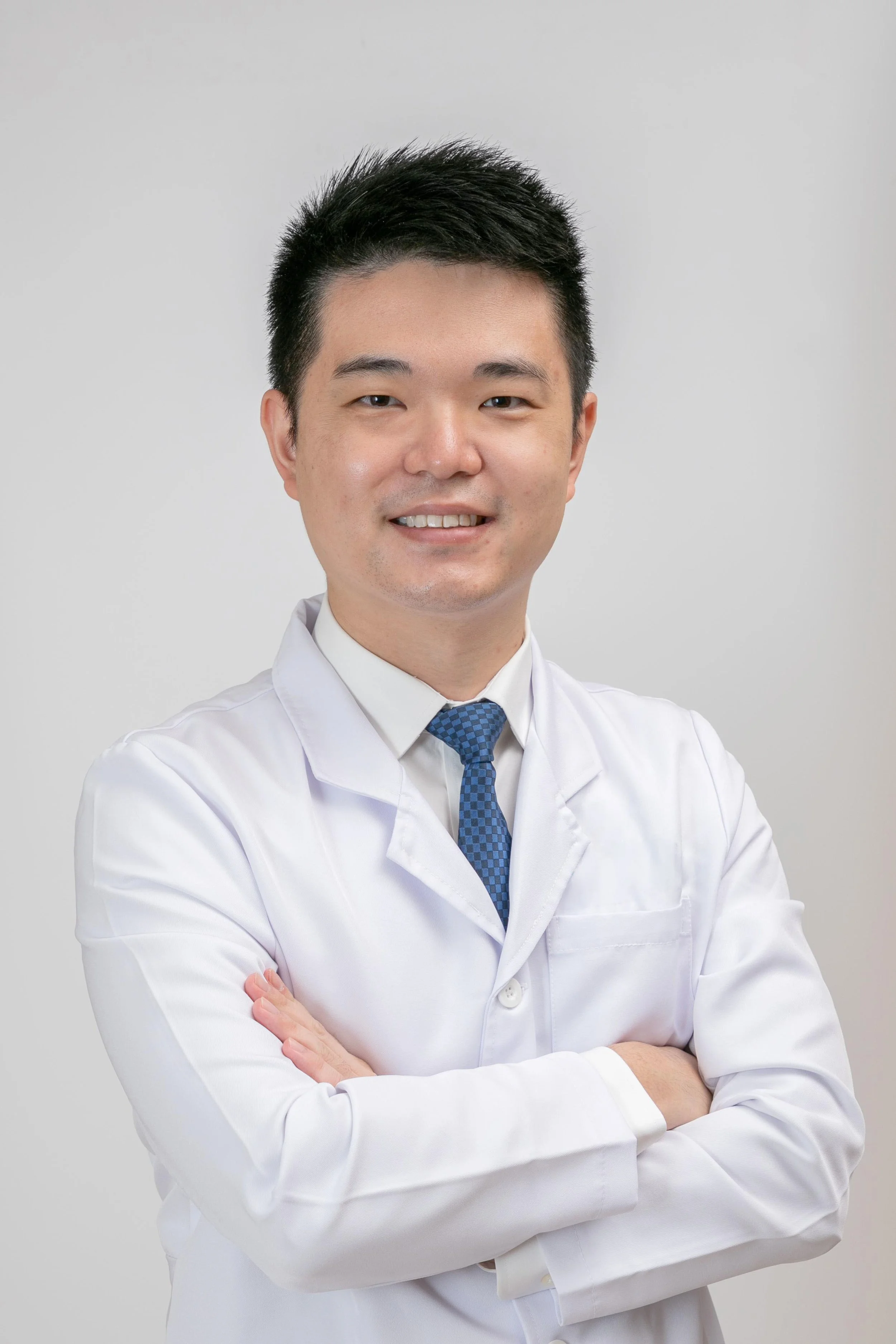 A male doctor with black hair, wearing a white lab coat and a blue tie, standing with arms crossed against a plain light gray background.