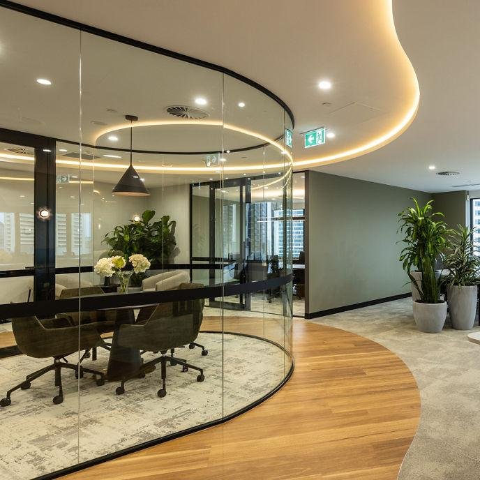 Modern office conference room with glass walls, black conference table, black chairs, and a floral centerpiece. The room is adjacent to an open lobby area with large potted plants, wooden flooring, and city views from windows.