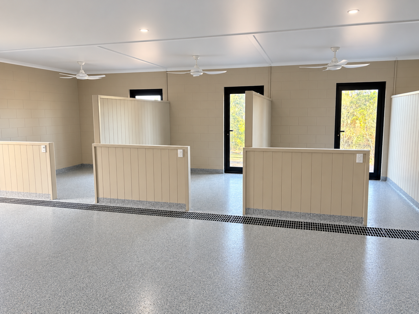Empty indoor facility with beige partitions, gray flooring, ceiling fans, and large windows
