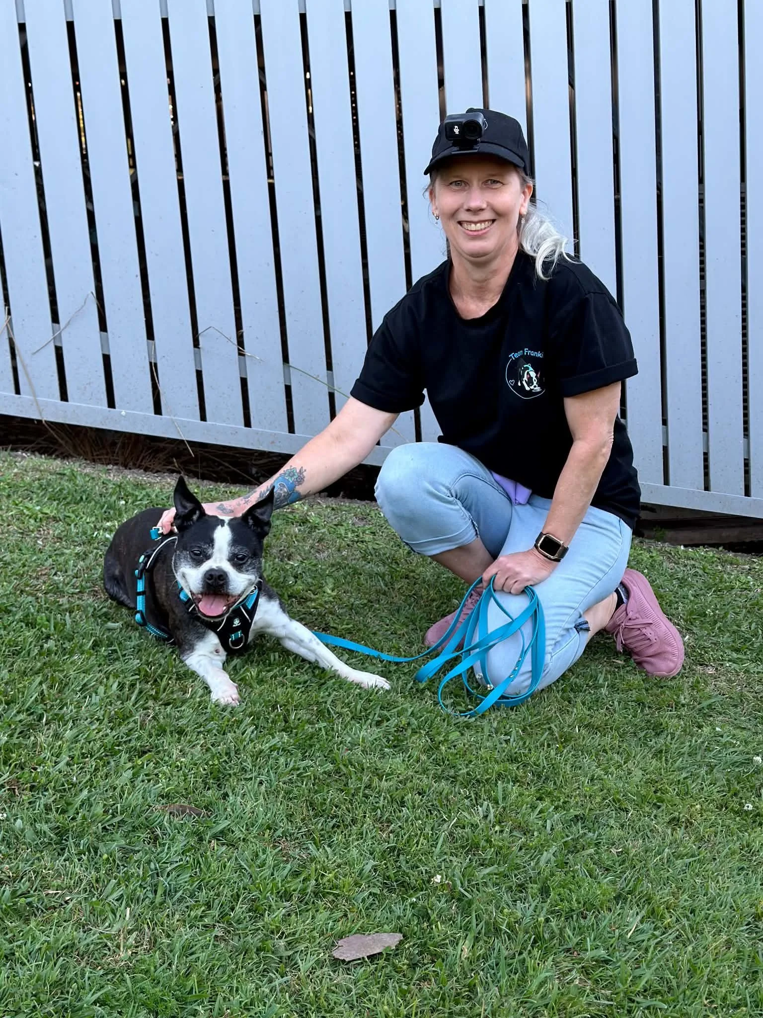 Smiling woman kneeling on grass beside a happy black-and-white dog wearing a blue harness and leash.