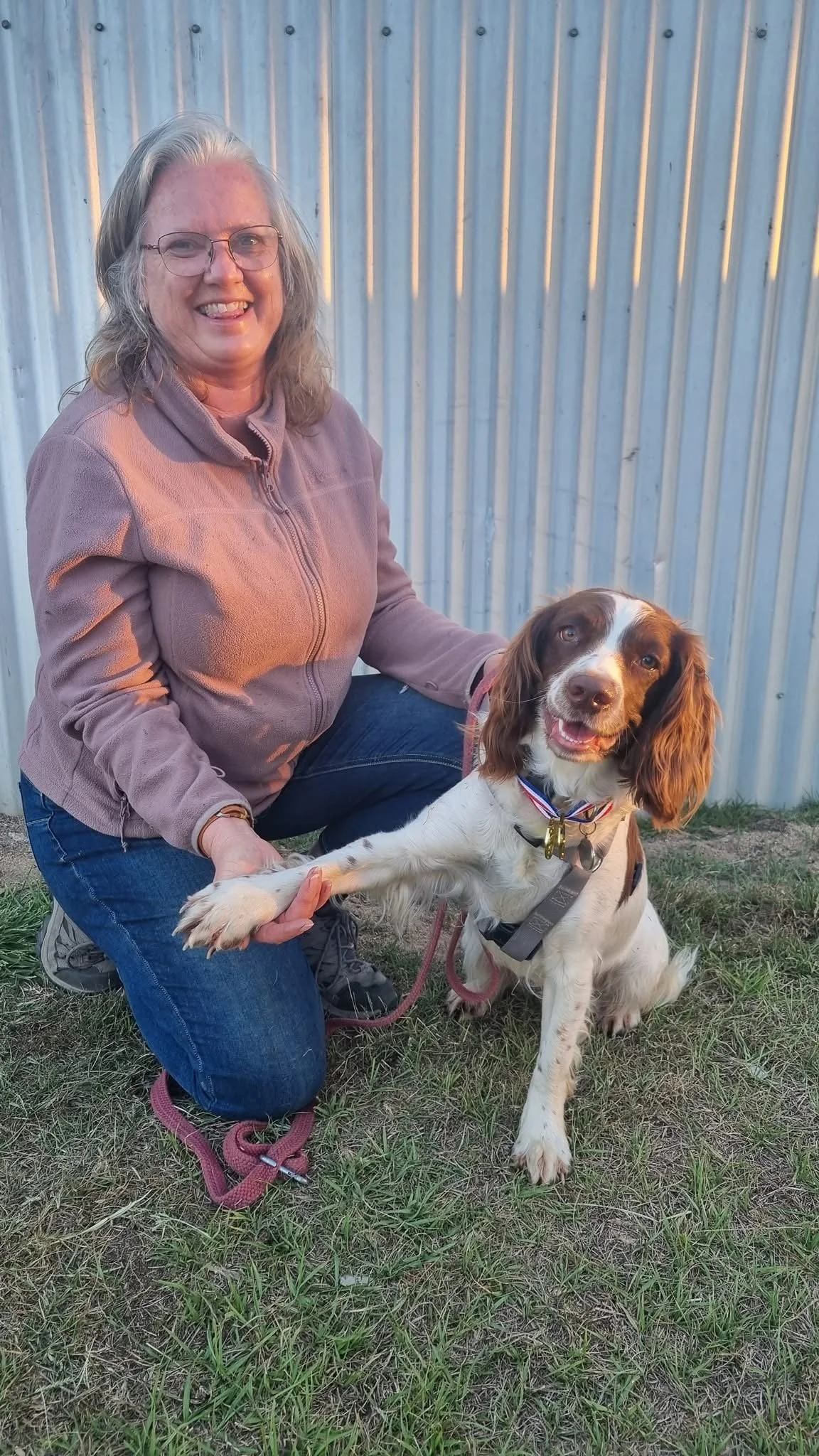 Smiling woman kneeling beside a medal-wearing brown-and-white spaniel outdoors