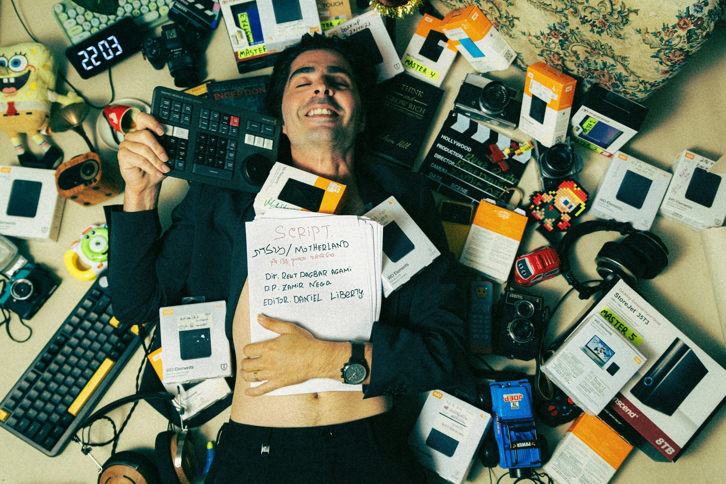 A man lying on a desk surrounded by tech gadgets, boxes of electronic products, and colorful items, smiling with eyes closed while holding a large keyboard in one hand and a script in the other.