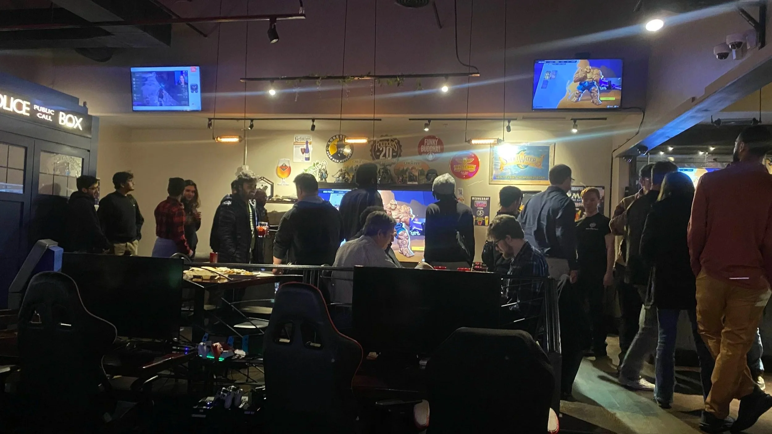 People gathered in a gaming bar or lounge watching screens showing video games, with some people playing and others socializing, and various posters and signs on the walls.